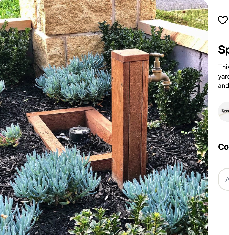 A garden sprinkler on a wooden post in a bed of succulent plants with dark mulch, near a stone wall and a white brick wall.