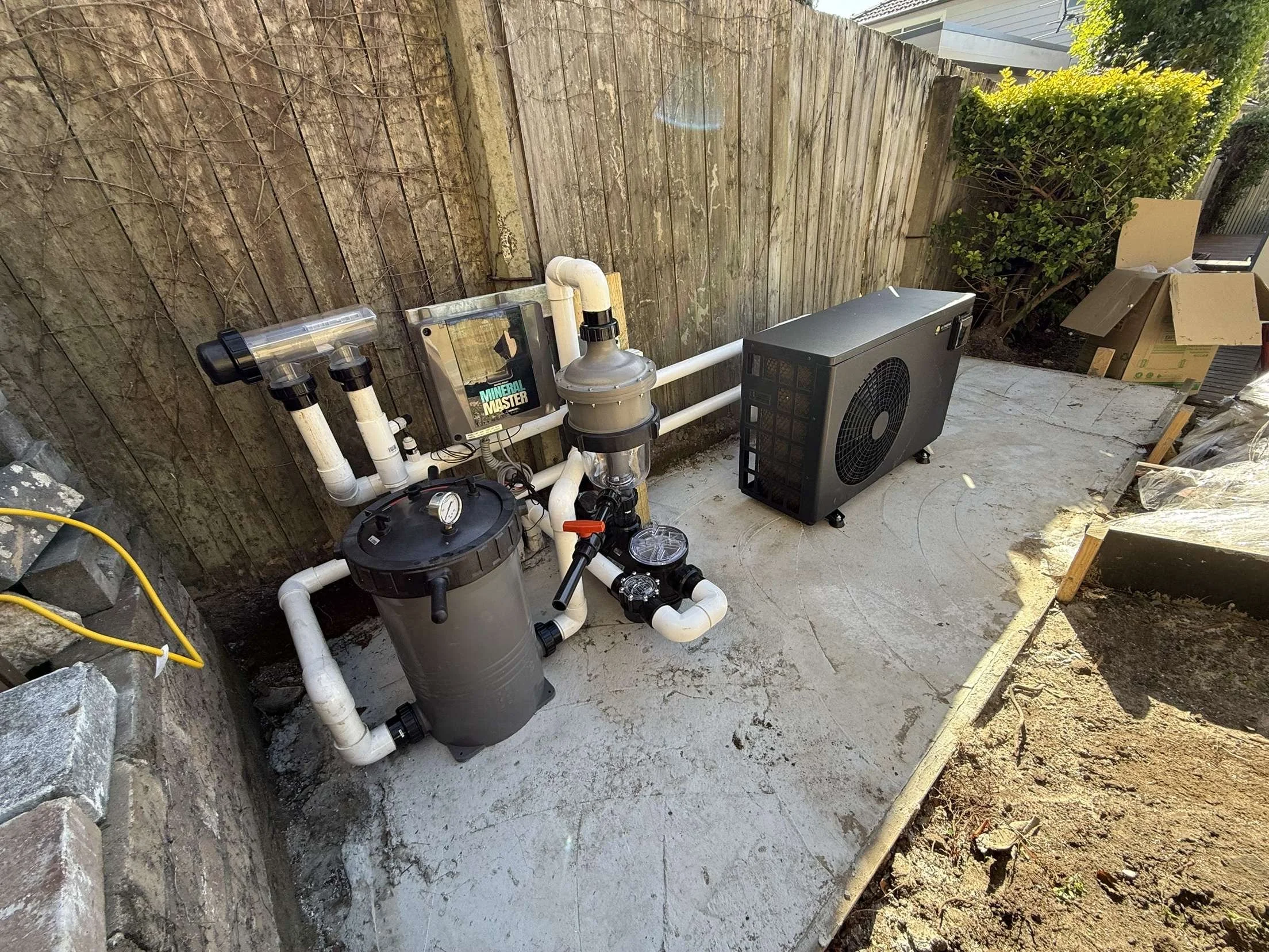Pool equipment including a pump, filter, cooler, and a chiller on a concrete pad outside a fenced backyard.
