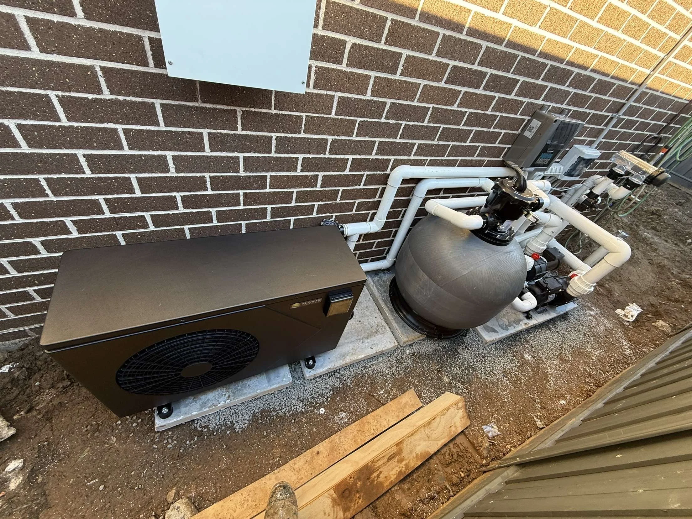Outdoor swimming pool equipment setup with a black pump and filter packed with white PVC pipes against a brick wall.