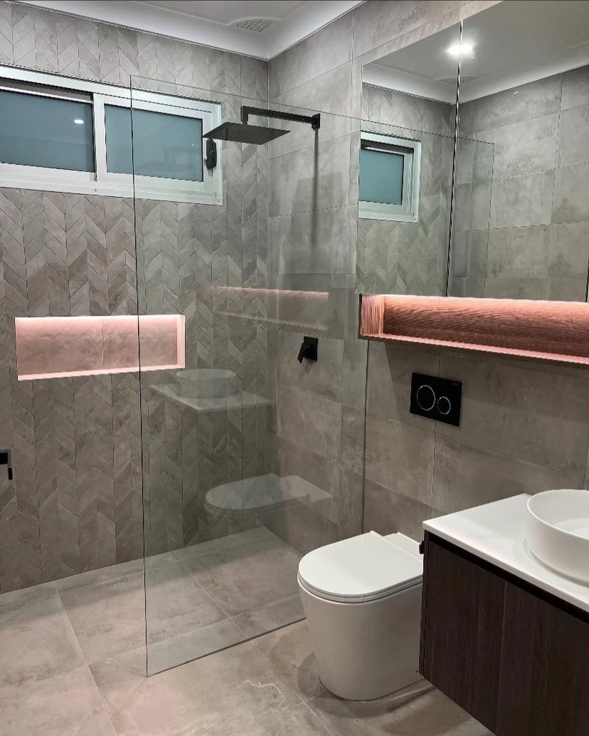 Modern bathroom with glass shower enclosure, wall-mounted toilet, dark wood vanity with vessel sink, and pink illuminated shelf, beige tiled walls, and two small horizontal windows.