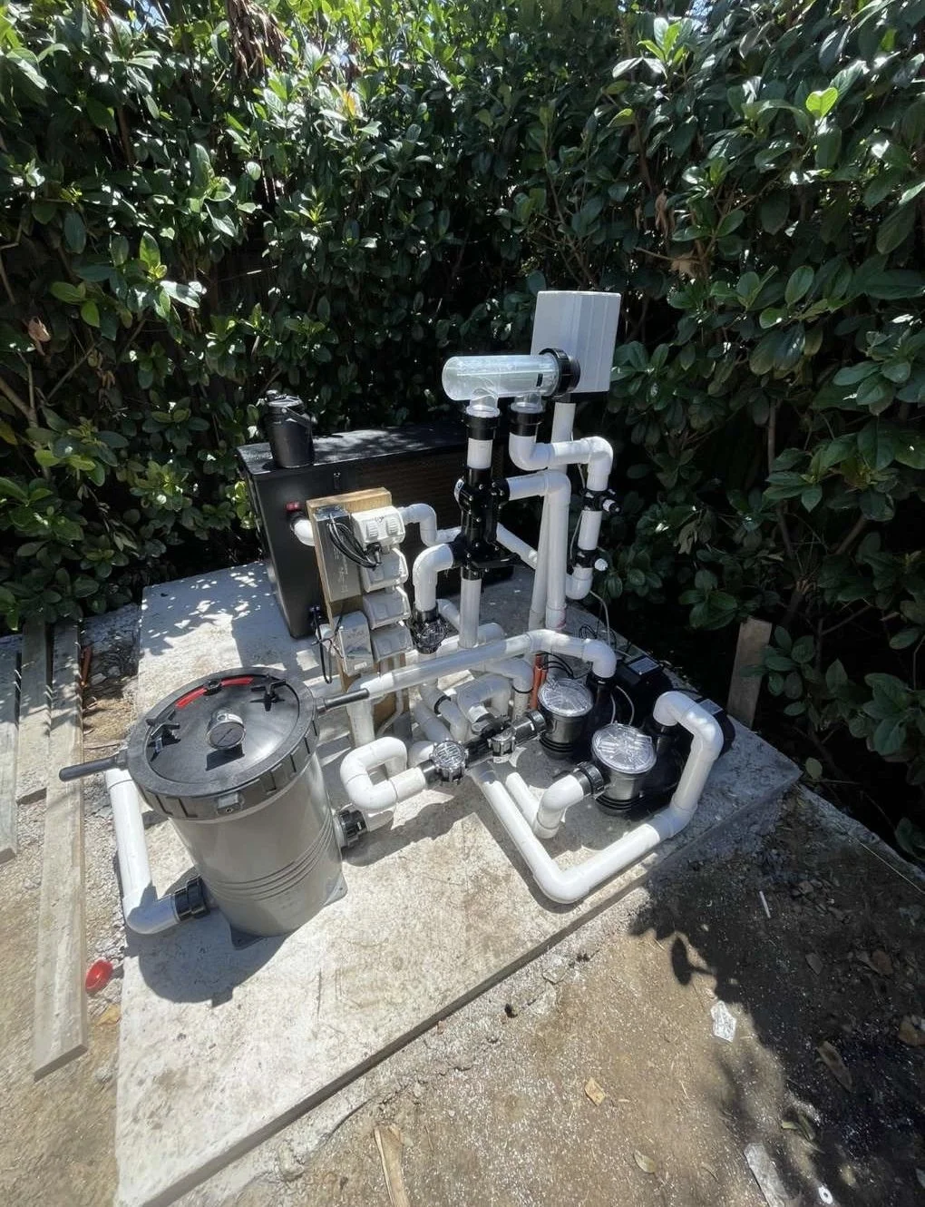 An outdoor pool filtration system with various white PVC pipes, pumps, and a sand filter, set on a concrete slab surrounded by green bushes.