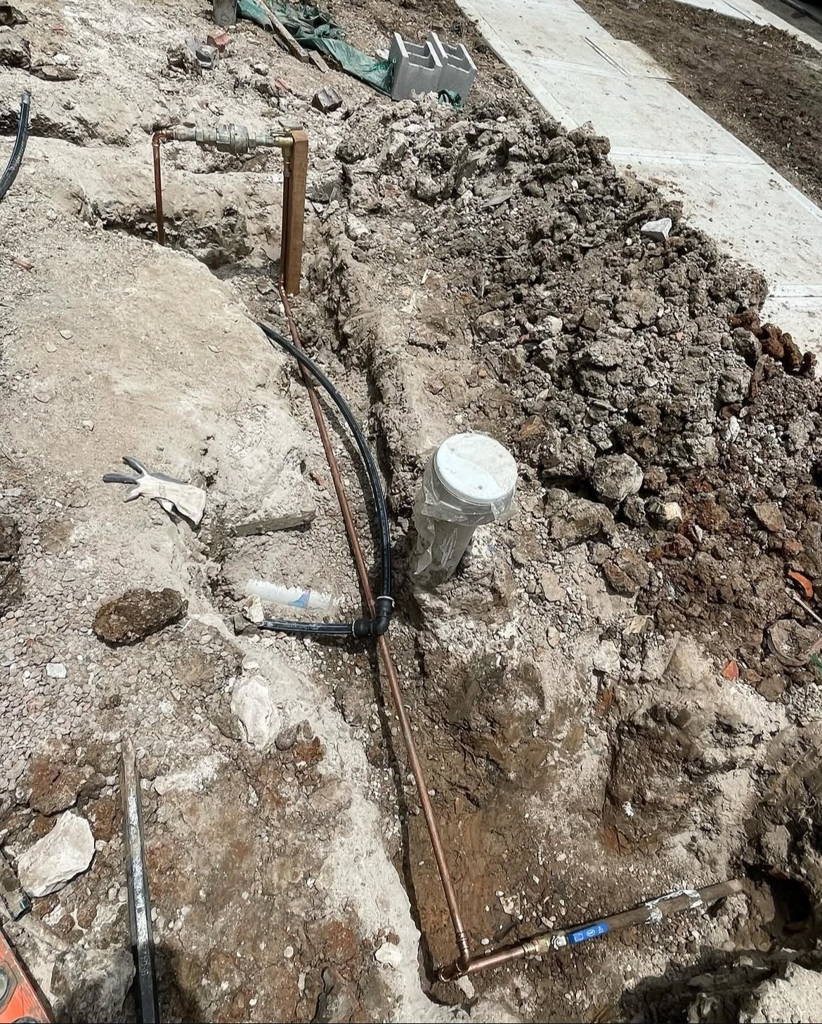 Underground plumbing installation with copper and black pipes in a dirt trench next to a concrete sidewalk.
