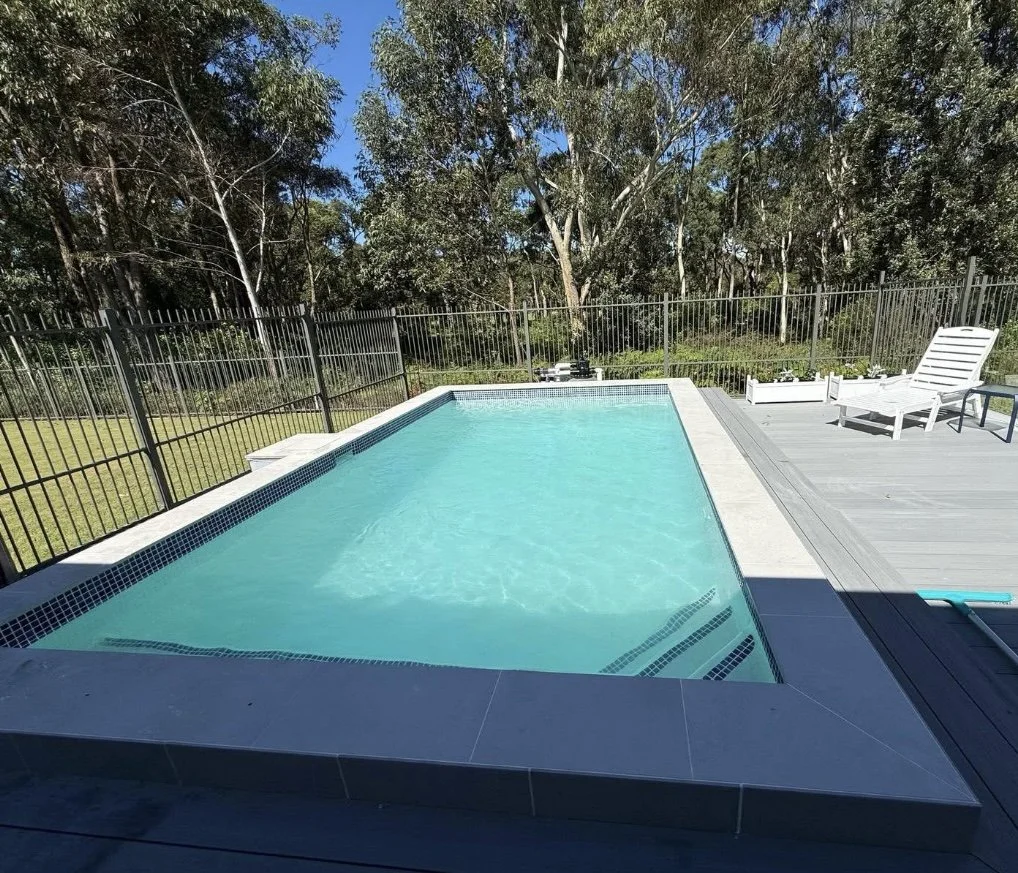 A backyard swimming pool with a black metal fence, outdoor lounge chair, and surrounded by trees on a sunny day.