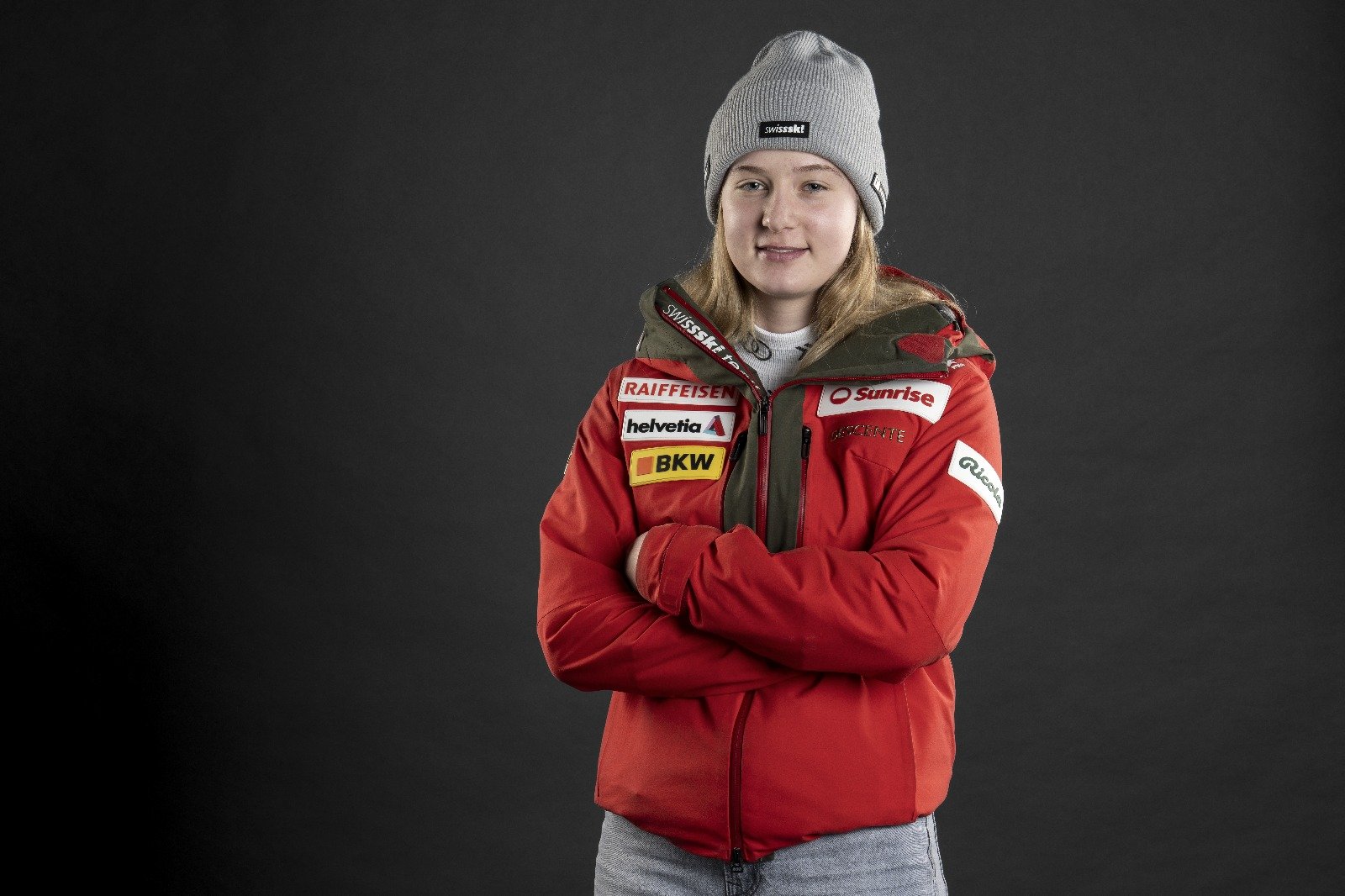 Young woman in a red jacket with sponsor patches, gray beanie, arms crossed, standing against a dark background.