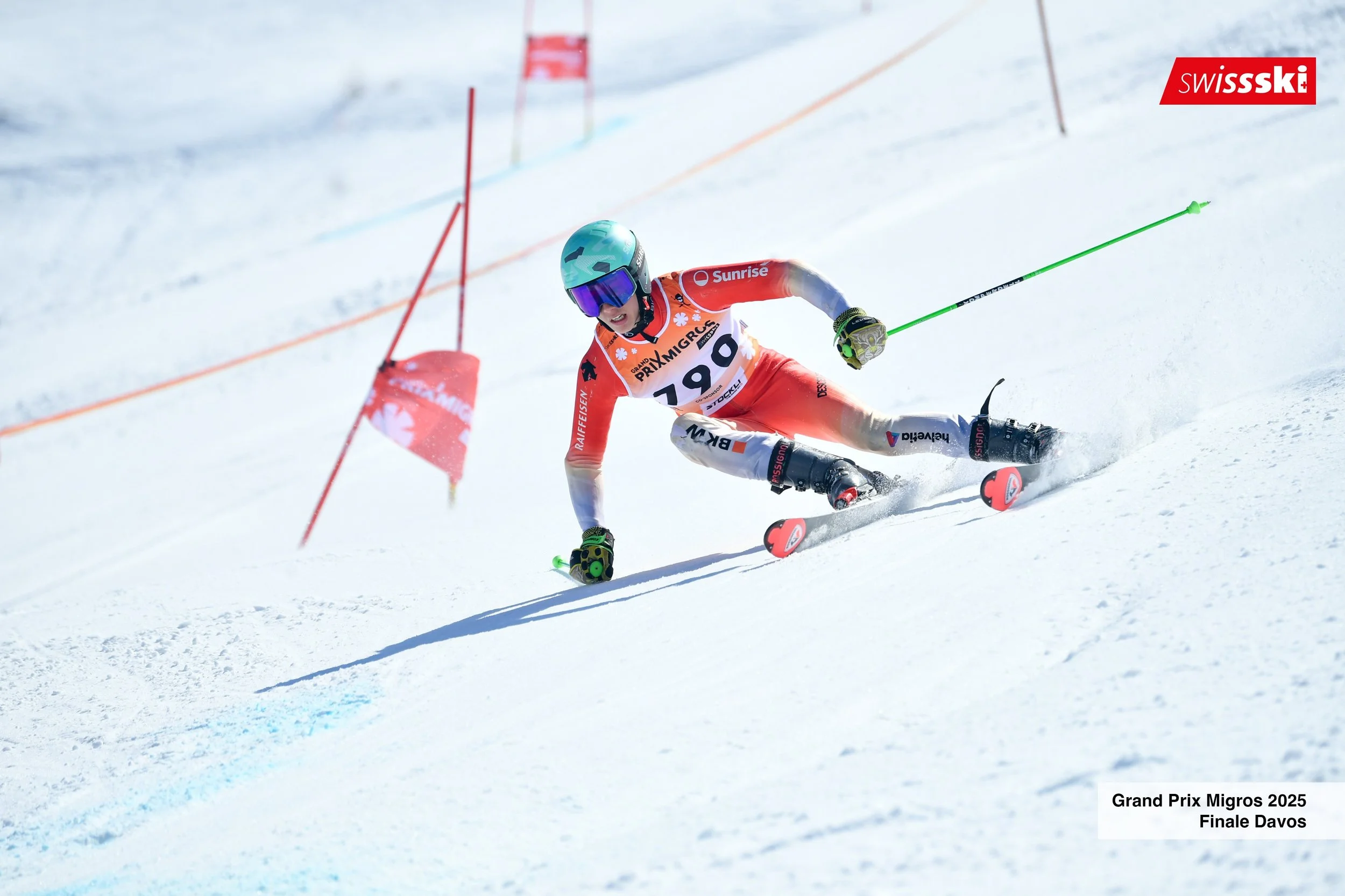 A skier in a red and white outfit with the number 1790 competes in a downhill race, making a turn on snow-covered slopes during the Grand Prix Migros 2025 Finals in Davos, with a red Swiss ski flag and orange safety netting in the background.