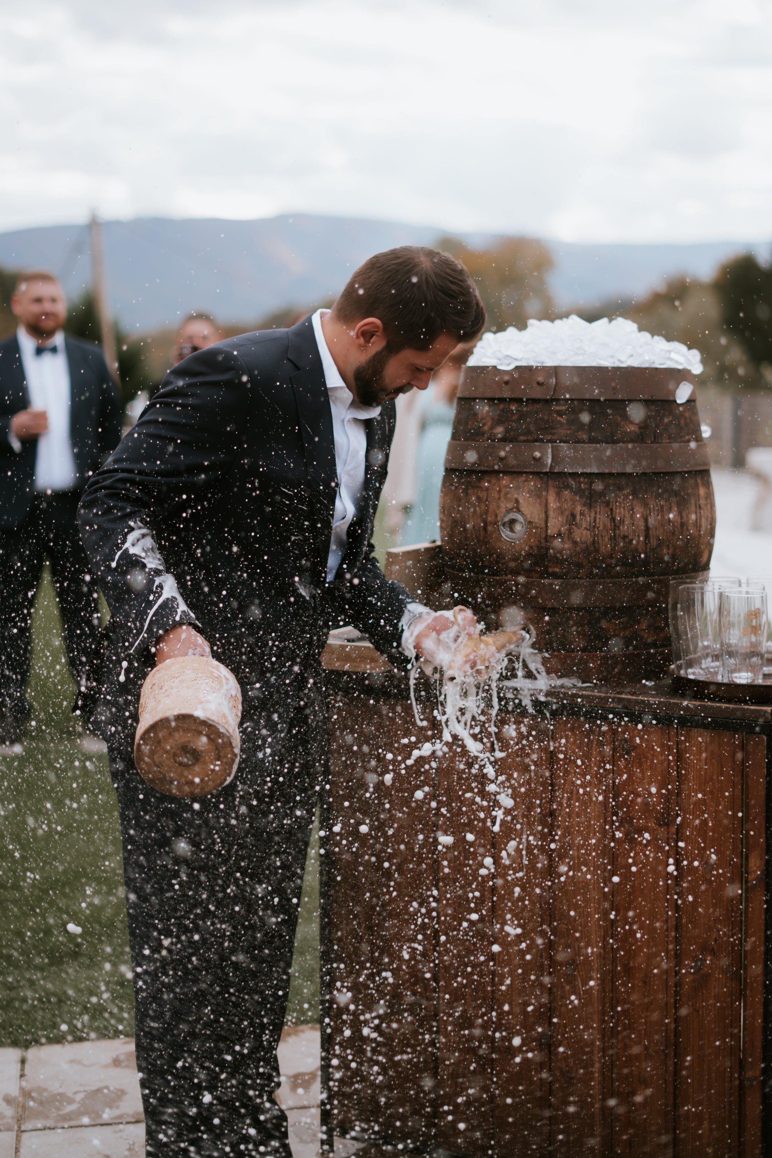 Man in tuxedo leaning over and slicing a cake with a hat, during a celebration outdoors with a wooden barrel and snow, with guests in tuxedos in the background.