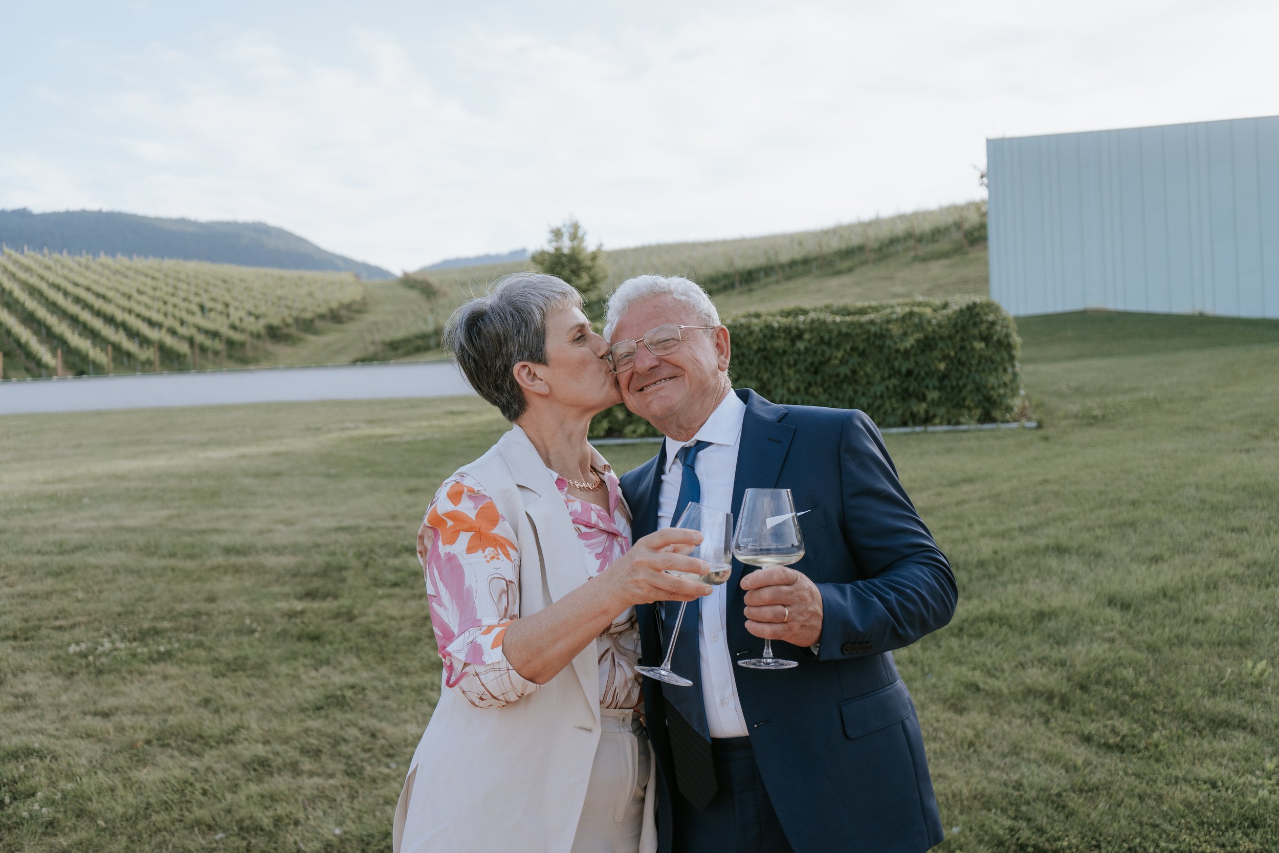 An elderly man and woman celebrating outdoors with wine glasses, kissing and smiling with a vineyard and mountains in the background.