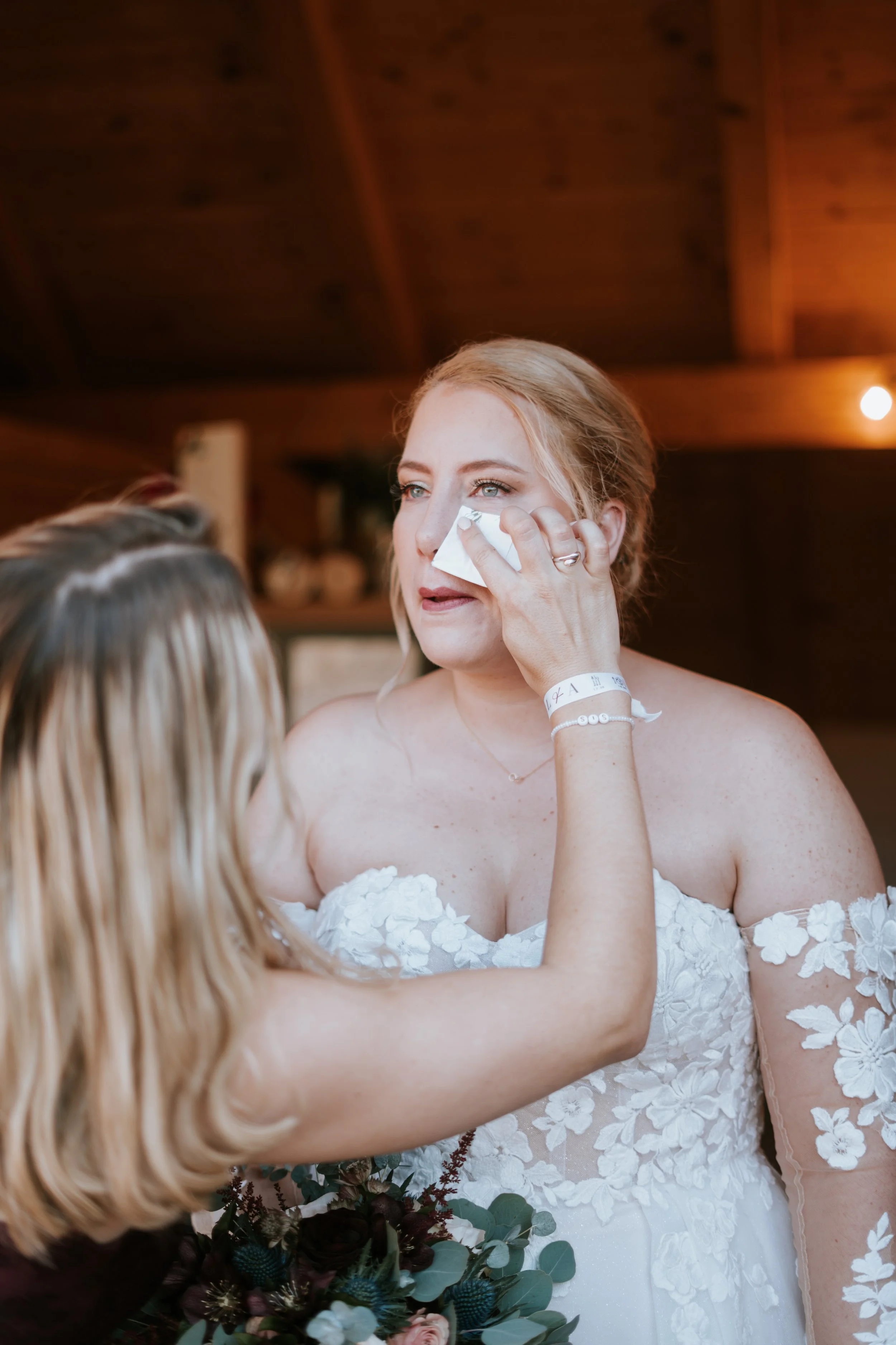 A woman with light blonde hair and blue eyes, wearing a white strapless dress with floral lace details, is wiping tears from her face with a tissue during a wedding celebration. She is holding a bouquet of dark purple, red, and green flowers with a m