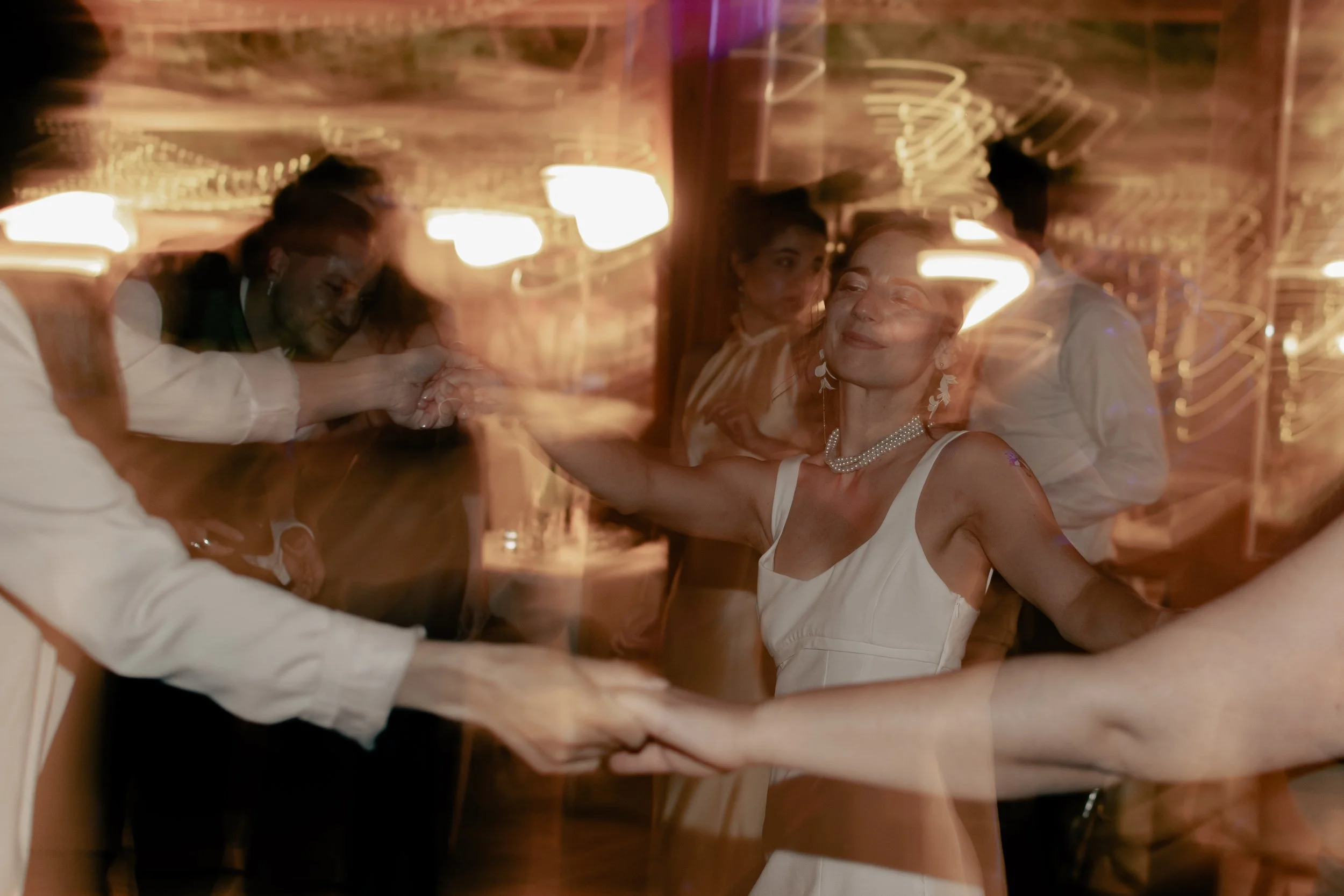 A woman in a white dress and pearl necklace dancing with a partner at a celebration or wedding, with blurry lights and other people dancing in the background.