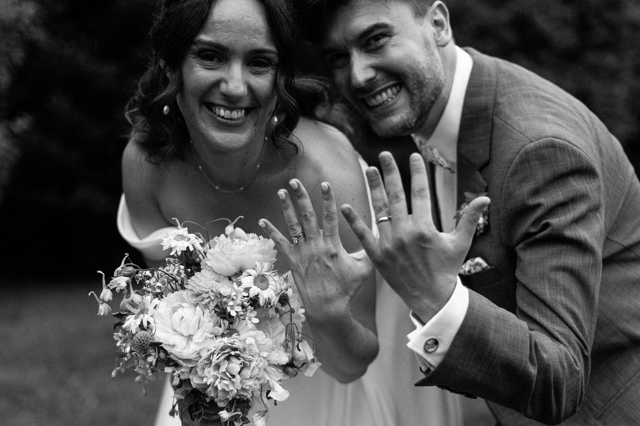 Close-up of a bride and groom with their foreheads and noses touching, smiling softly, under a bridal veil with white floral details, outdoors during daytime.
