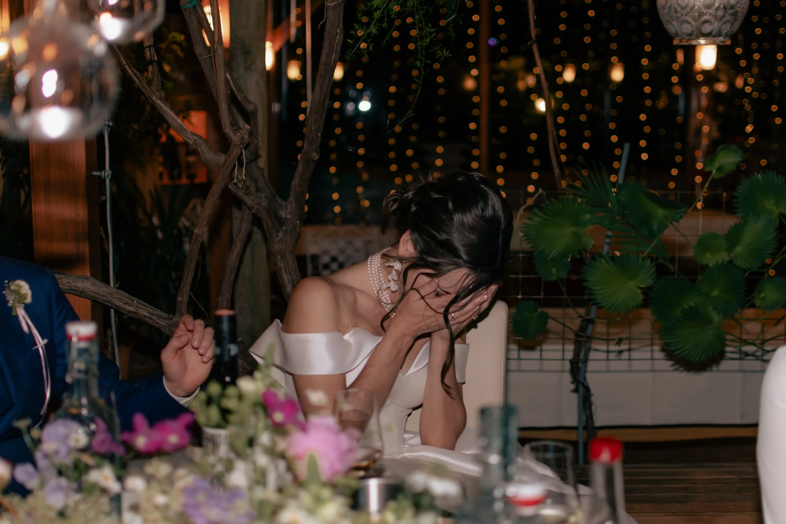 Woman in white dress with shoulder cutouts, wearing a pearl necklace, covering her face with her hands, sitting at a table with floral centerpiece and other guests at a dimly lit event.