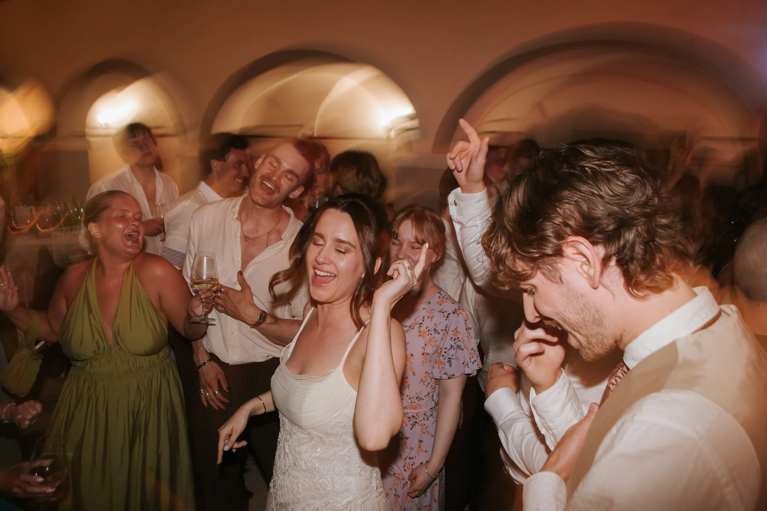 Group of people dancing and enjoying themselves at a party or celebration, with some individuals laughing and smiling, and colorful lighting creating a lively atmosphere.