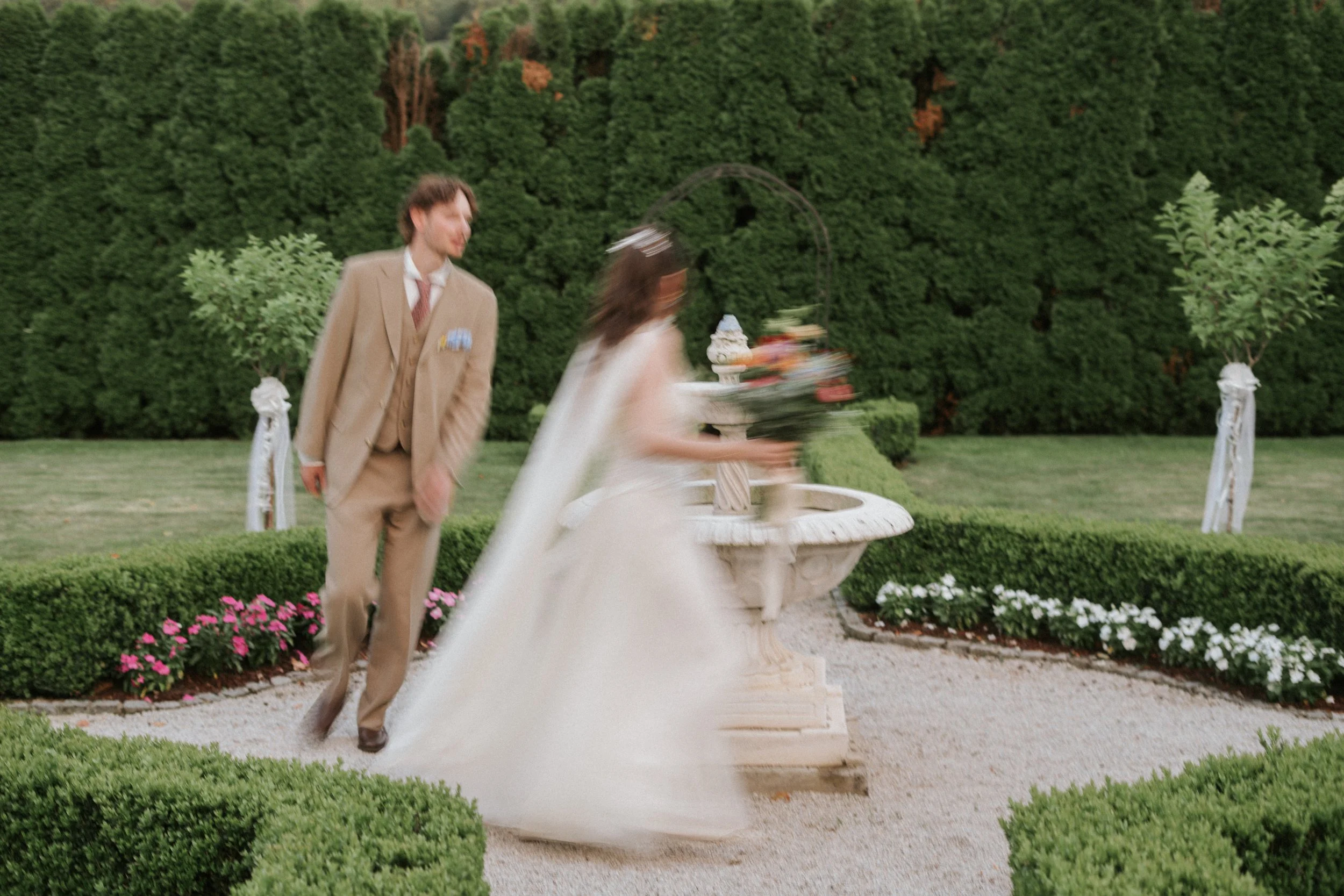 A man in a tan suit walking towards a bride in a white wedding dress, both are outdoors near a fountain with a lush green hedge in the background.