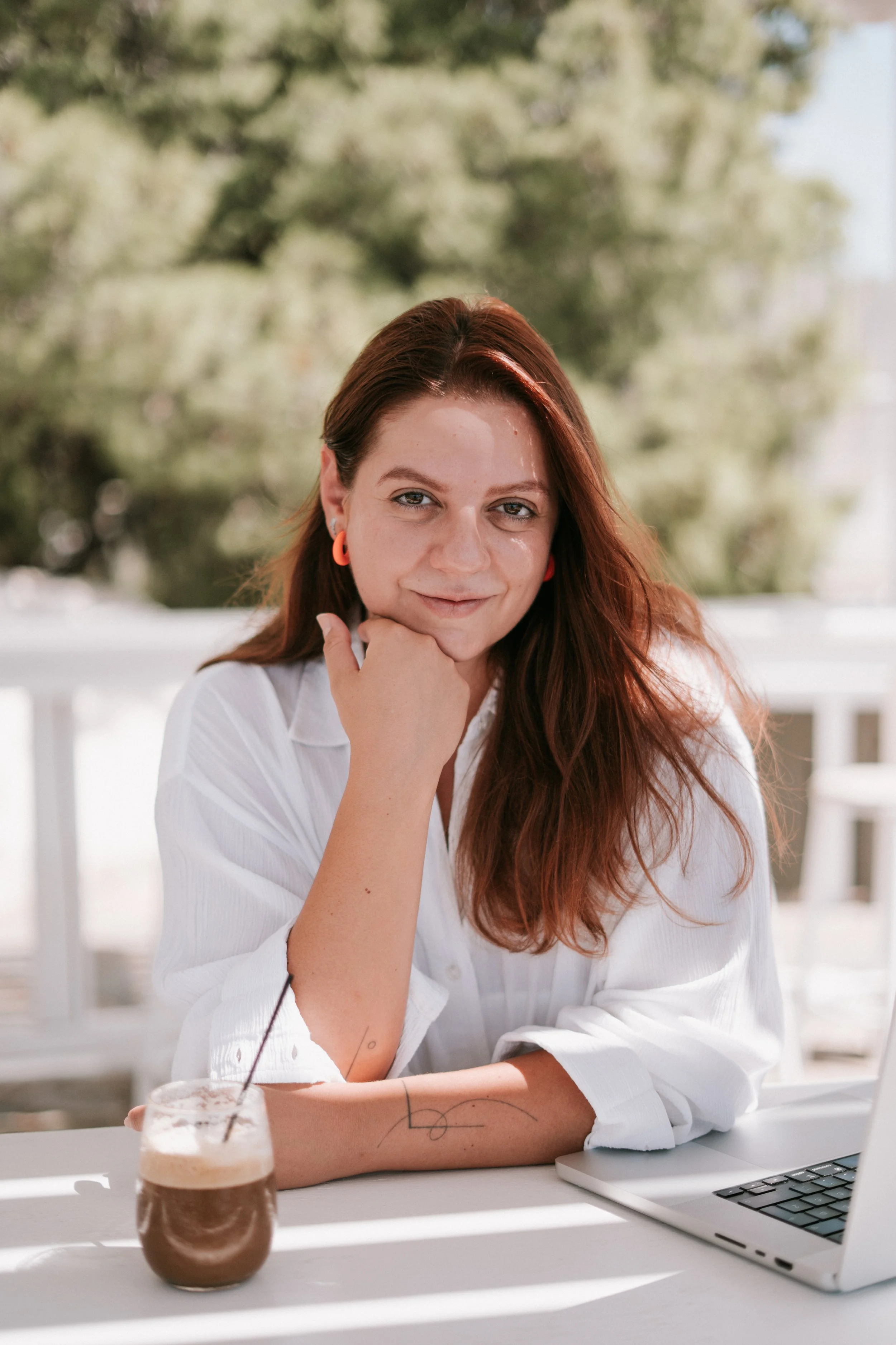 Hristina Trajkoska the photographer behind HTR studio with red hair and orange earrings looks at the camera, resting her chin on her hand, sitting at a table with a laptop and a drink outdoors.