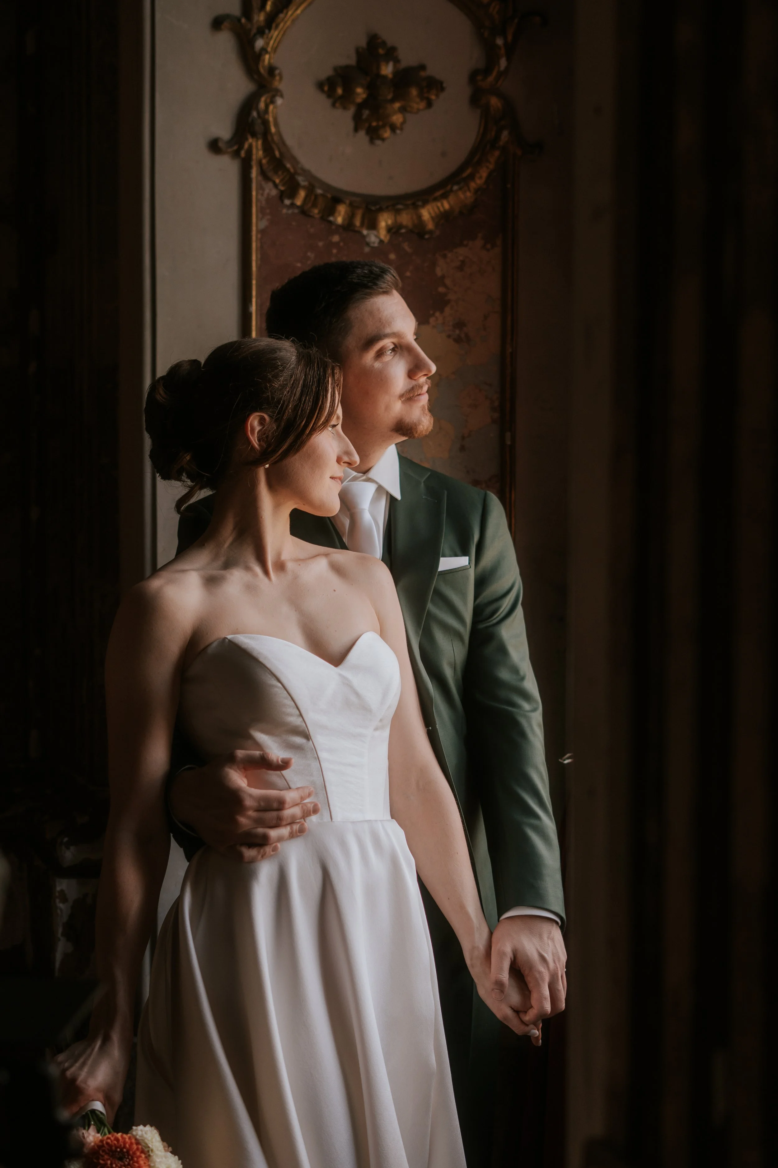 A bride and groom holding hands with closed eyes, standing close, indoors with ornate wall decor in the background.