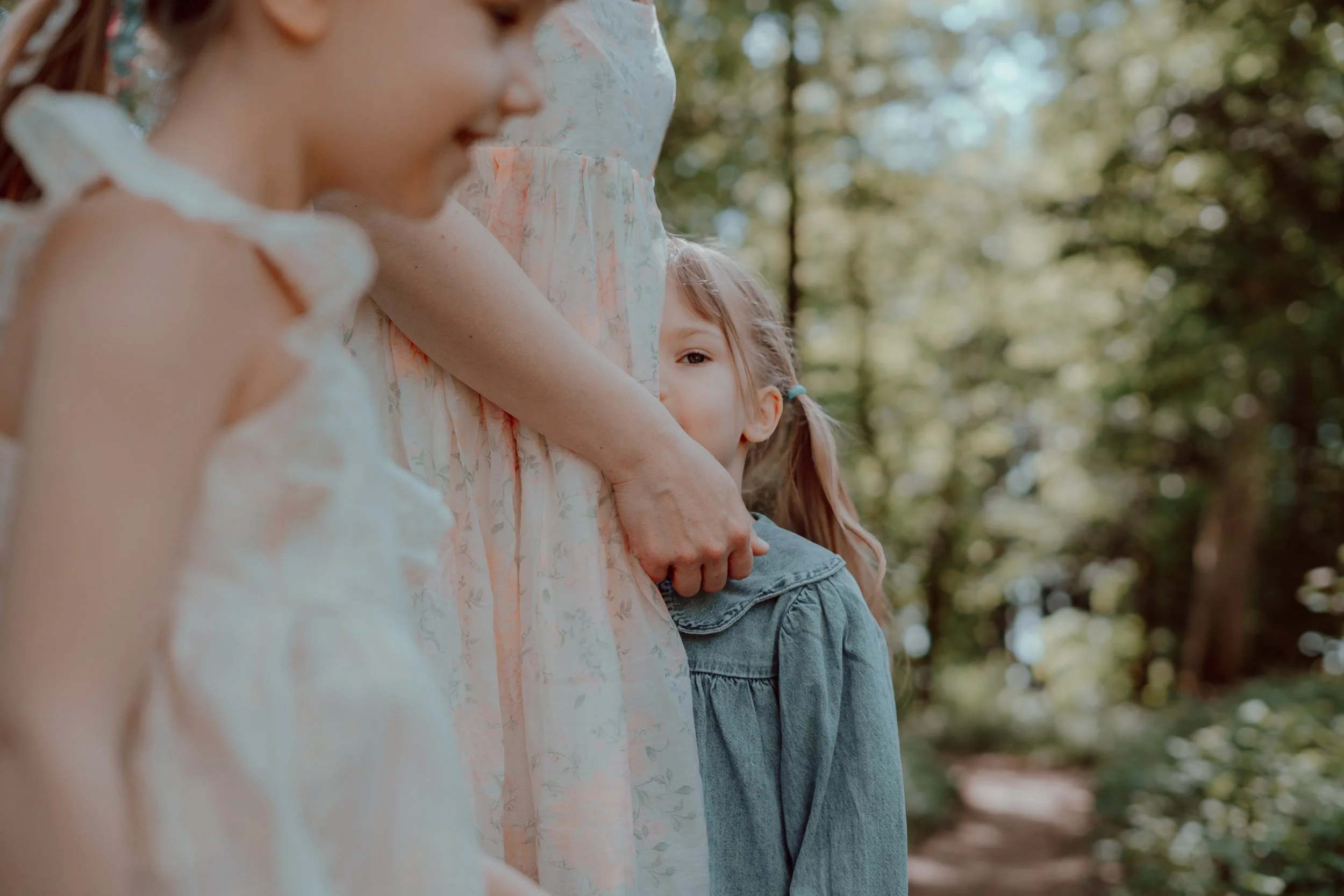 A woman with two young girls in an outdoor wooded area, with one girl hiding behind the woman, peeking over her arm, and the other girl smiling softly.