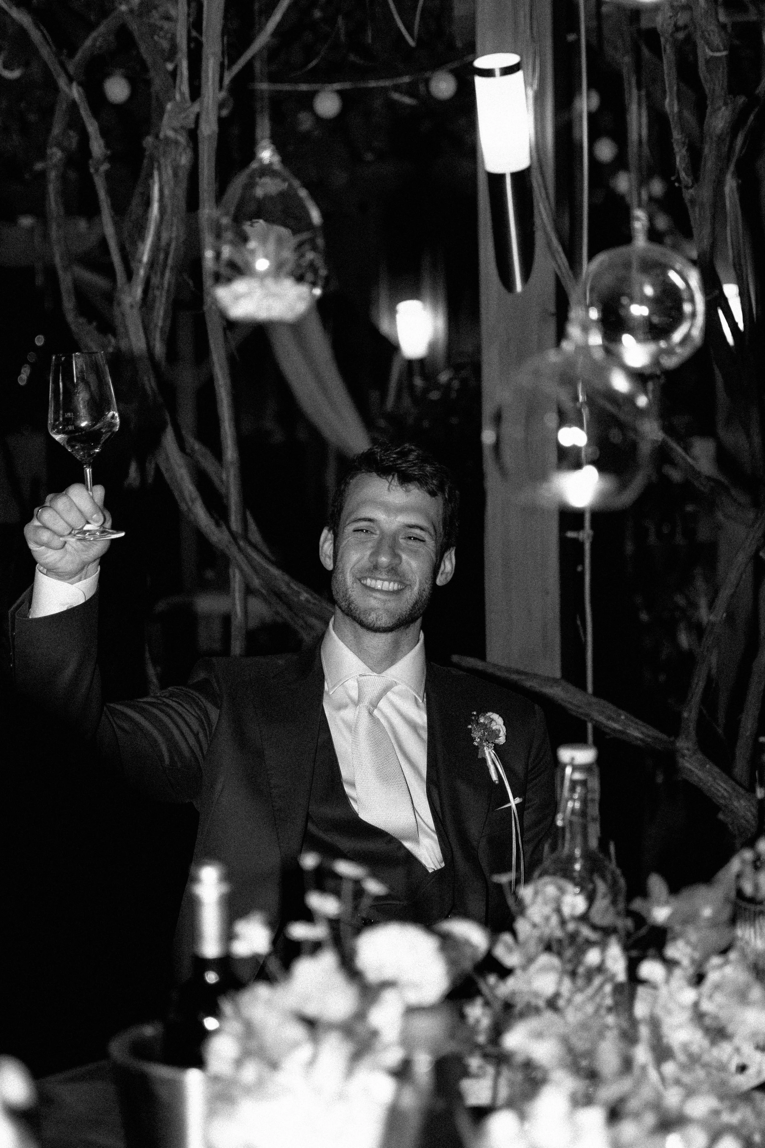 A man in a tuxedo smiling and raising a glass at a celebration or wedding reception, with decorative hanging ornaments and floral arrangements in the background.