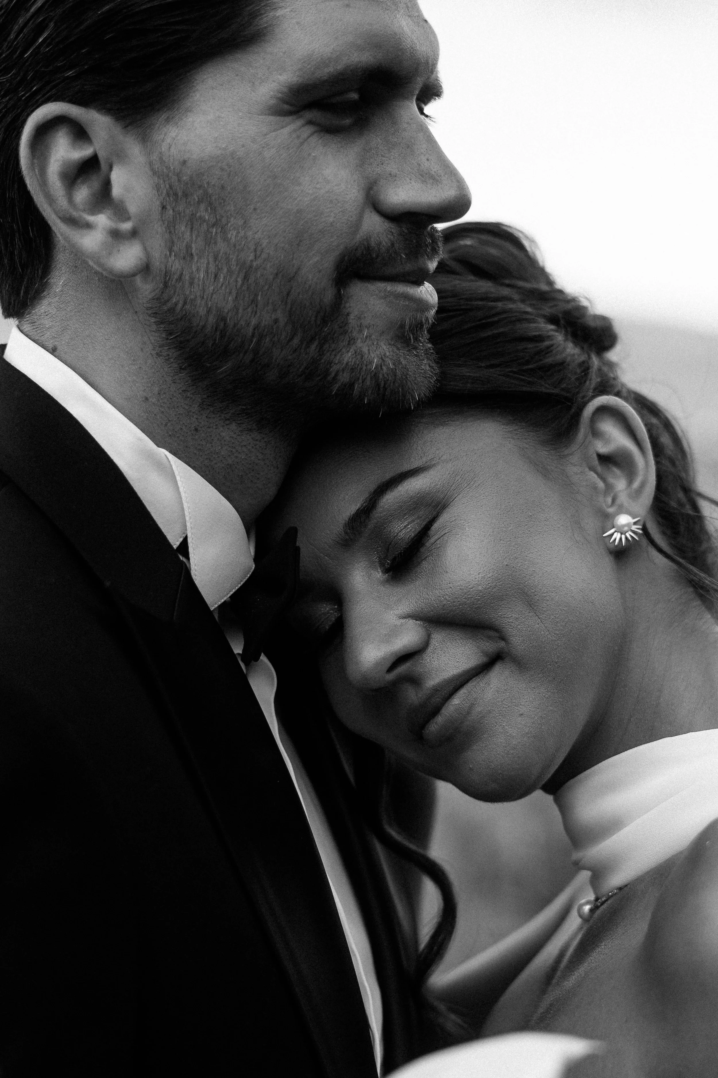 Black and white photo of a man in a tuxedo with a beard, smiling, and a woman with closed eyes, a flower-shaped earring, and a smile, leaning her head on his shoulder.