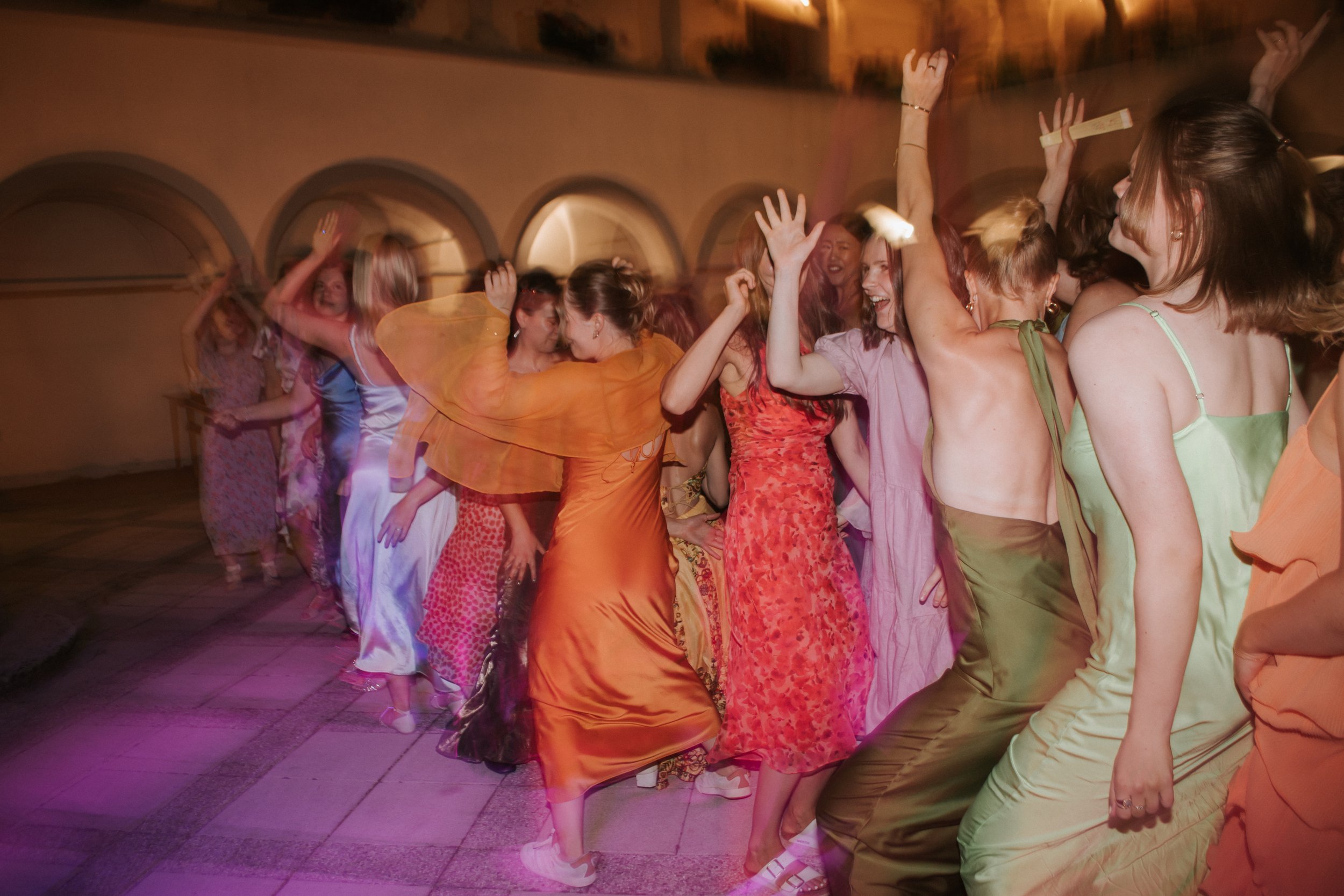 Group of women dancing and celebrating at an indoor party or event, wearing colorful dresses.