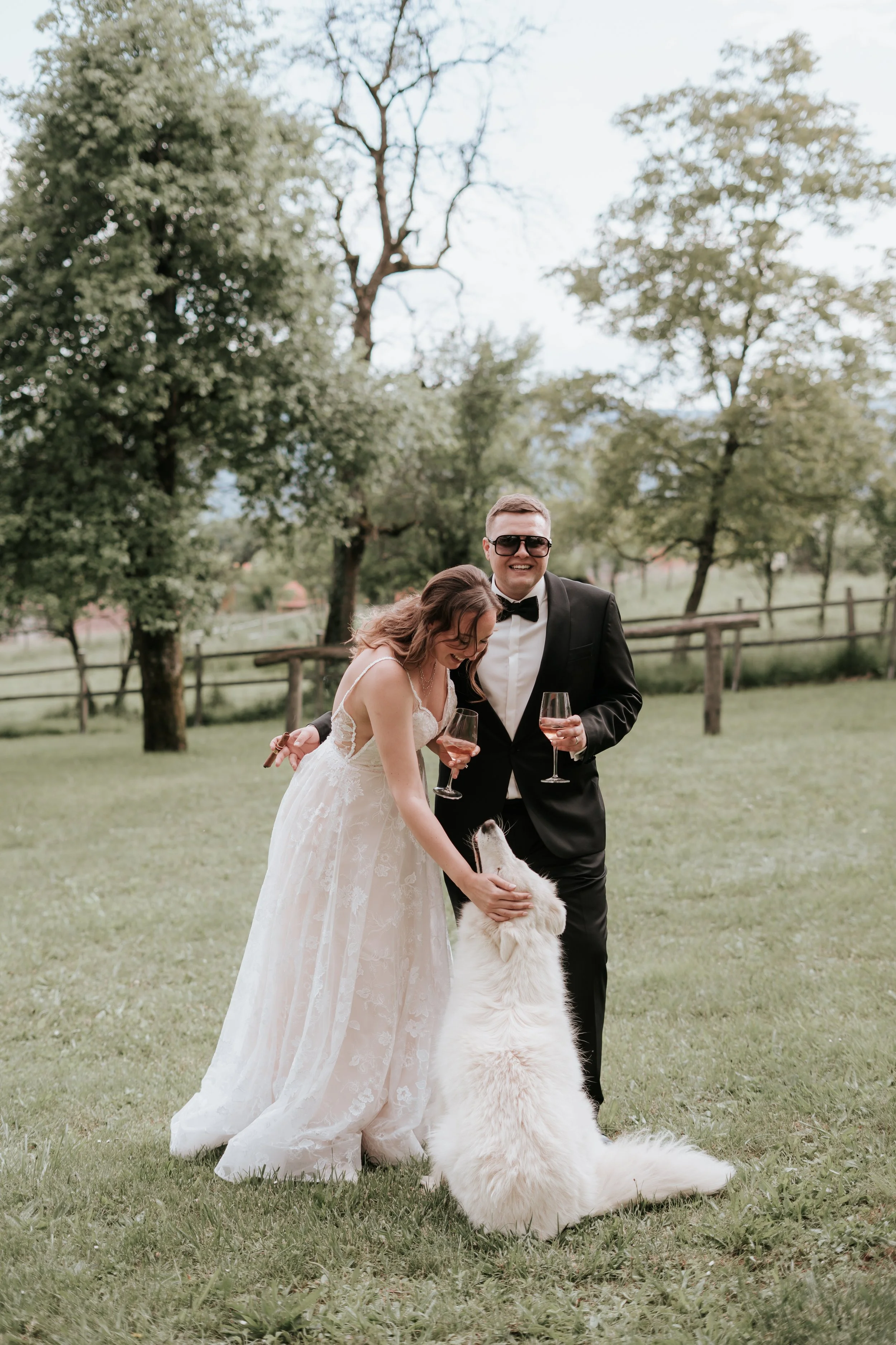 A bride and groom at a wedding reception outdoors, holding glasses of wine, petting a large, fluffy white dog on a grassy field with trees and a wooden fence in the background.