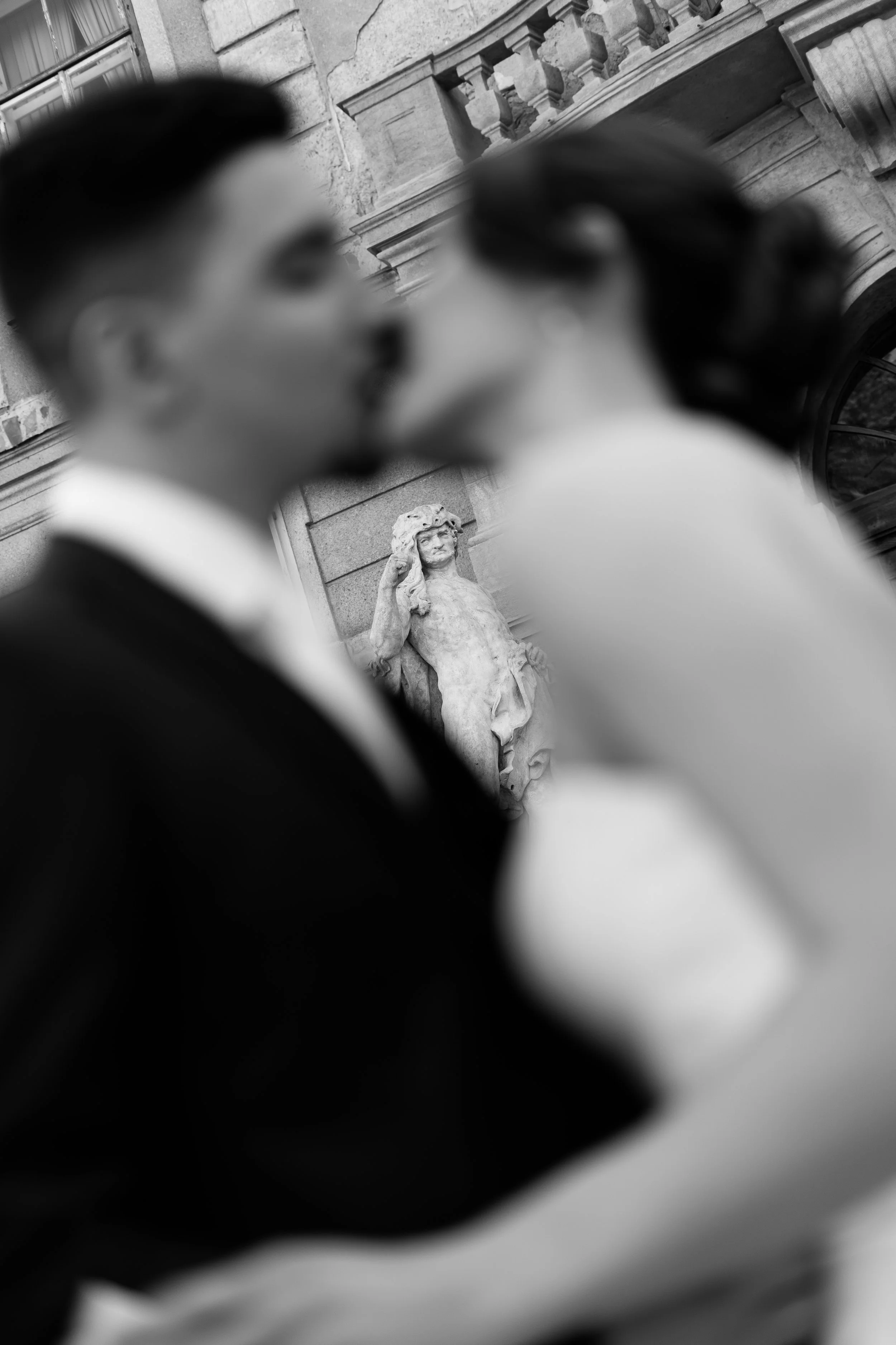 Black and white photo of a couple kissing, with a stone building and a statue of a woman in the background.
