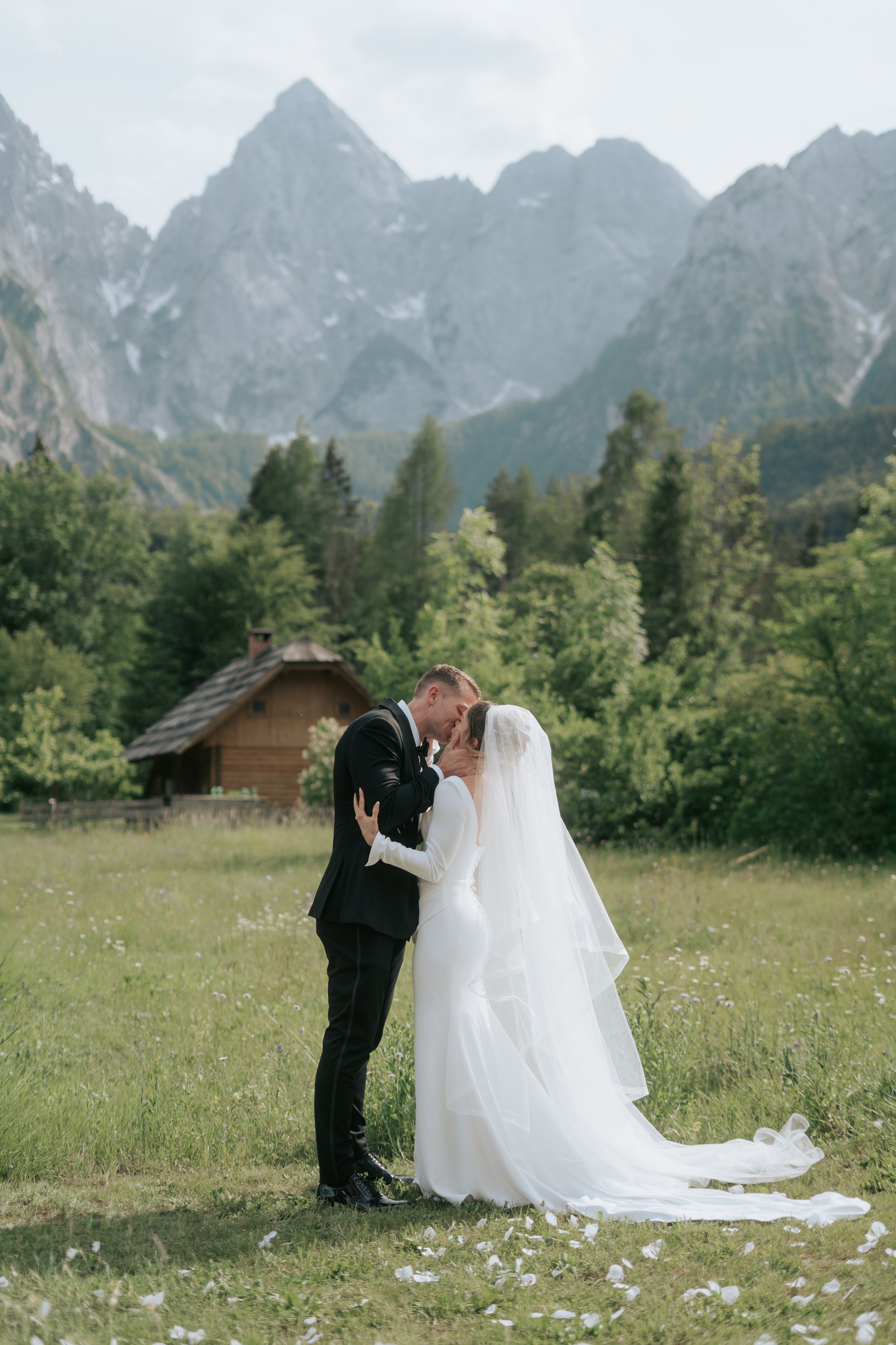 Close-up of a bride and groom with their foreheads and noses touching, smiling softly, under a bridal veil with white floral details, outdoors during daytime.