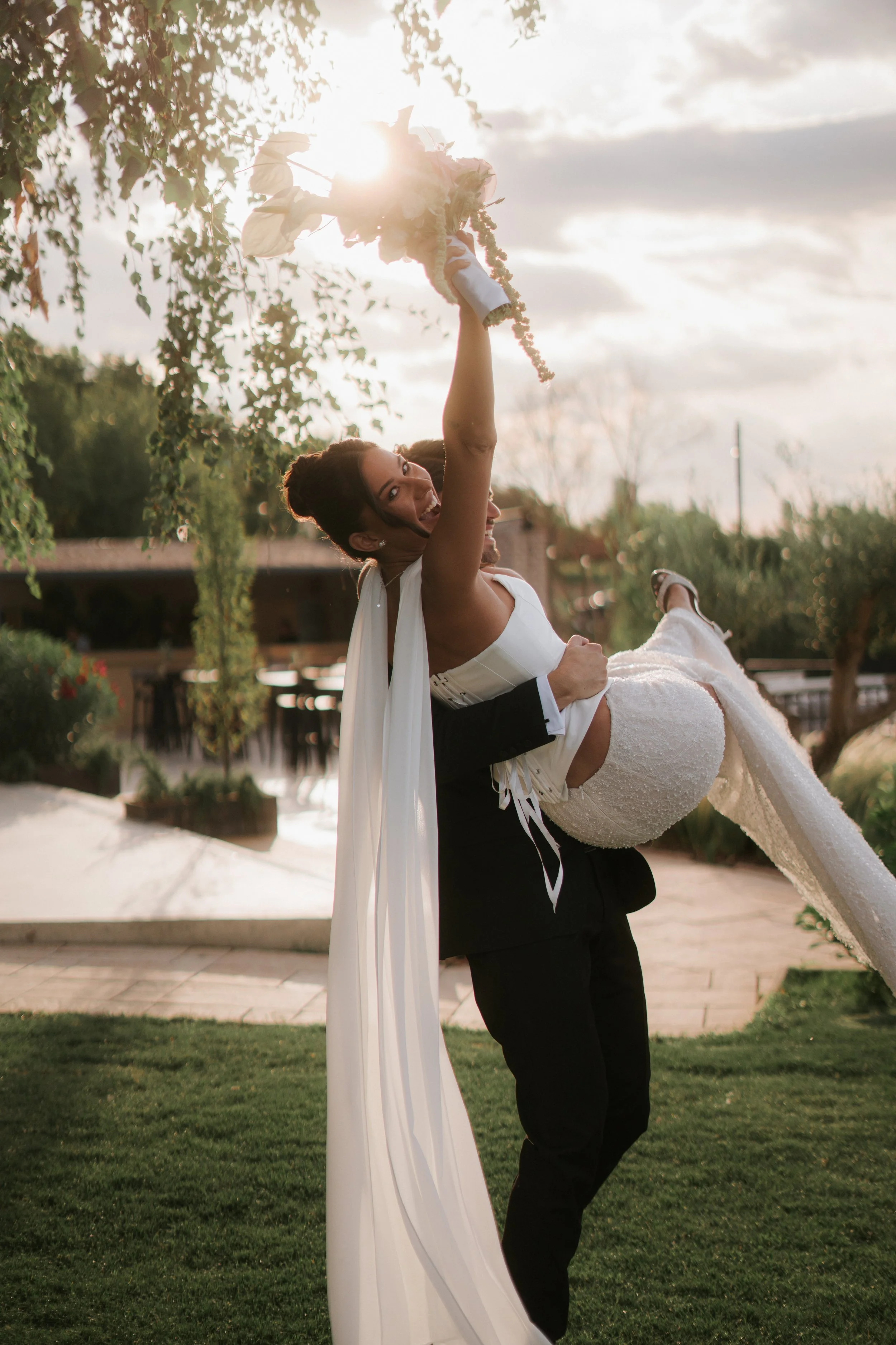 A bride and groom outdoors at sunset, with the groom lifting the bride while holding a bouquet. The bride is smiling and wearing a white wedding dress, and the groom is in a black suit.