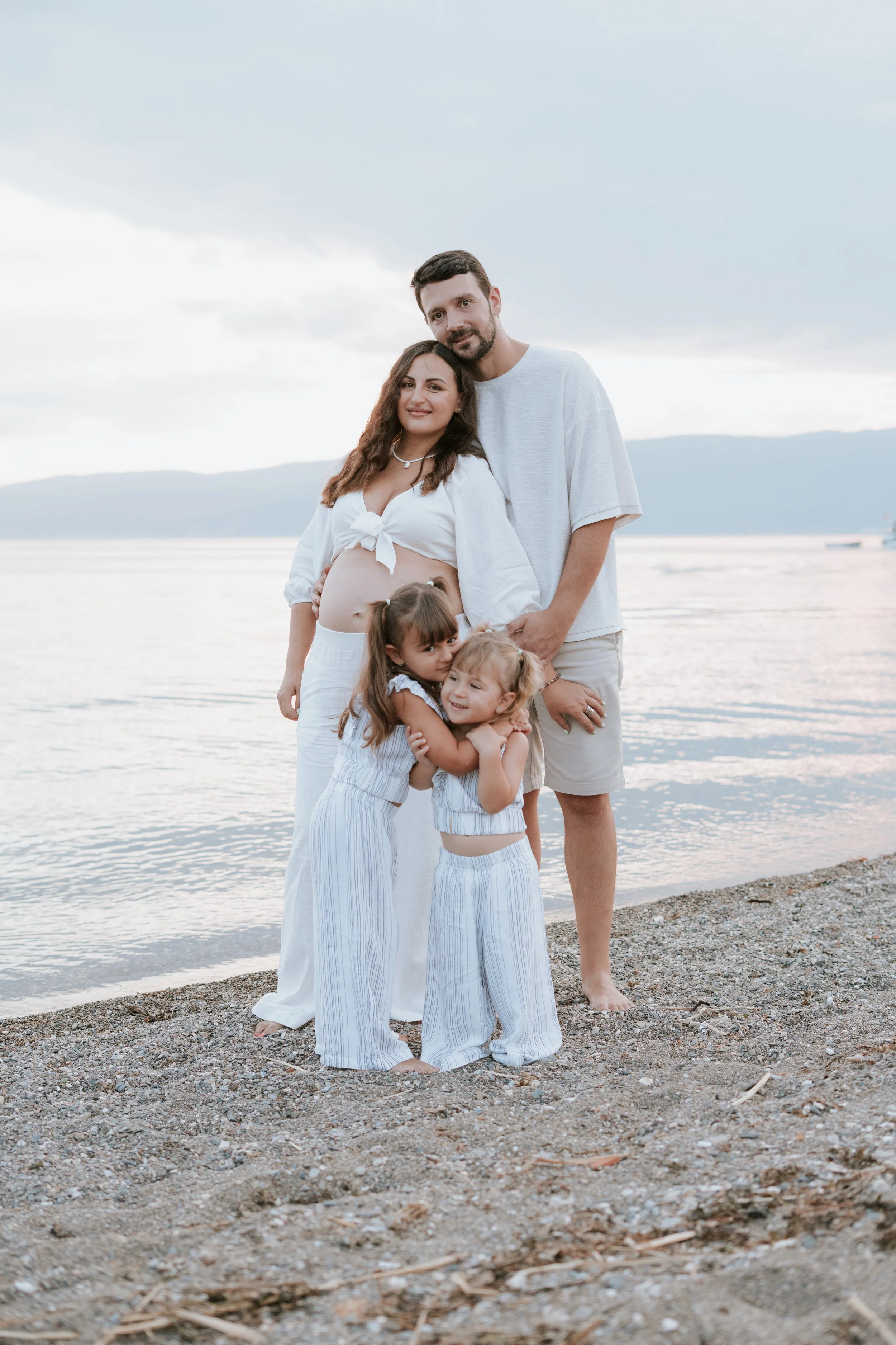 A family of four standing on a beach near water, with a cloudy sky and distant mountains in the background. The mother is pregnant, wearing white, and the father is beside her. Two young girls, hugging, stand in front of them, all dressed in light-co