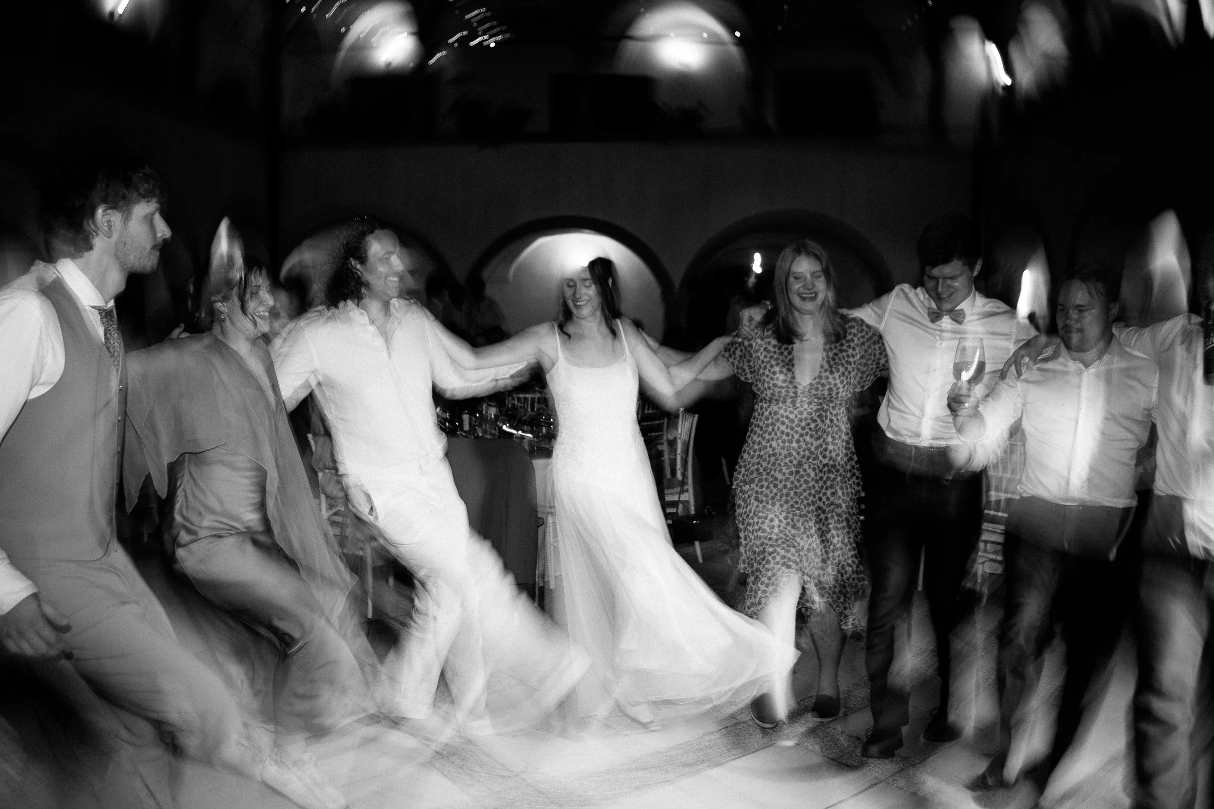 Group of people dancing in a circle at a party or celebration, smiling and holding hands, in a dimly lit room.