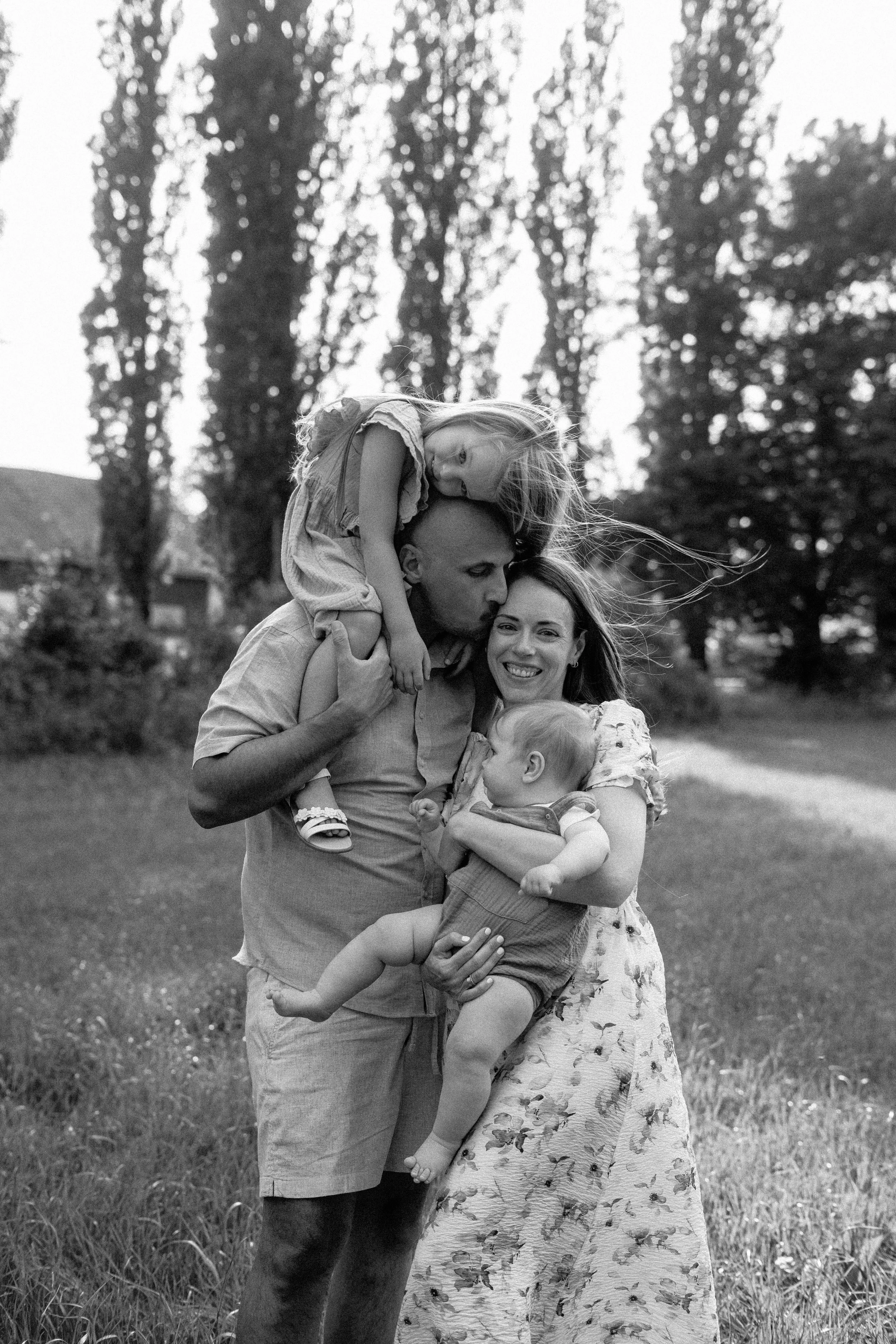 A black and white photo of a family outdoors, with a man holding a young girl on his shoulders, a woman holding a toddler, and a young boy in front. The girl is leaning over the man's head, and the boy is looking up at her. The woman is smiling, and they are surrounded by tall trees and grass.