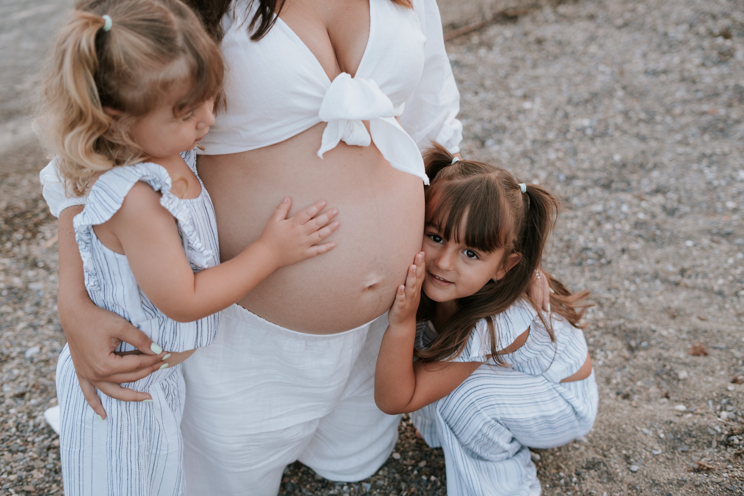 A pregnant woman with two young girls, one touching her belly and the other leaning her head against her belly, outdoors on a gravel surface.
