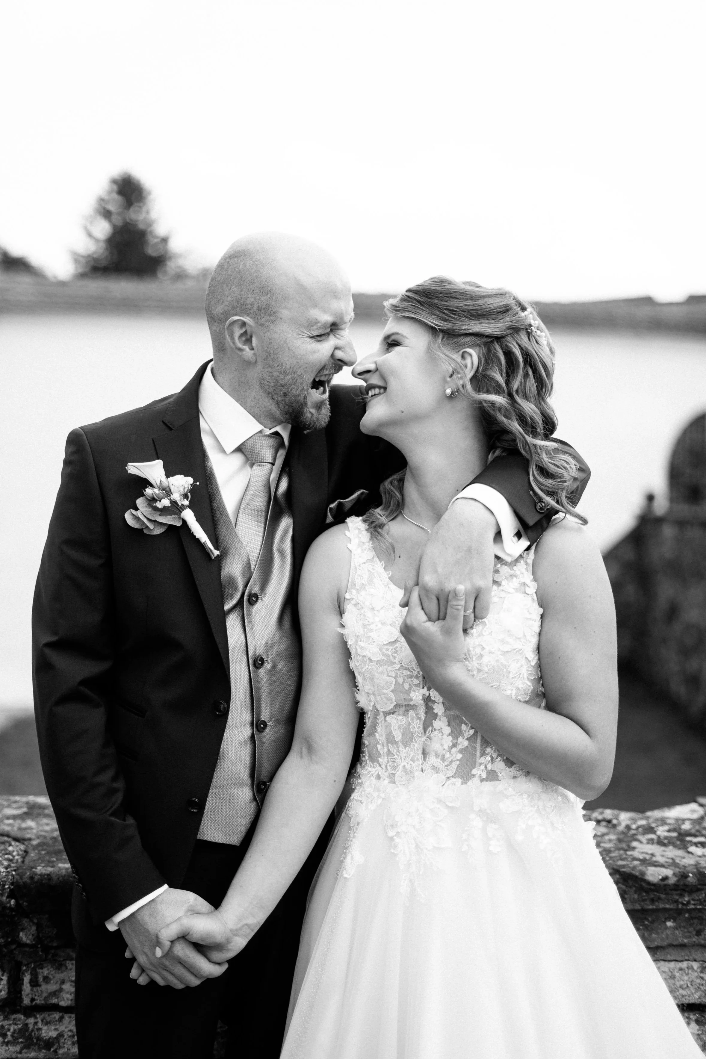 A couple hugging by the water during sunset, smiling and embracing each other.