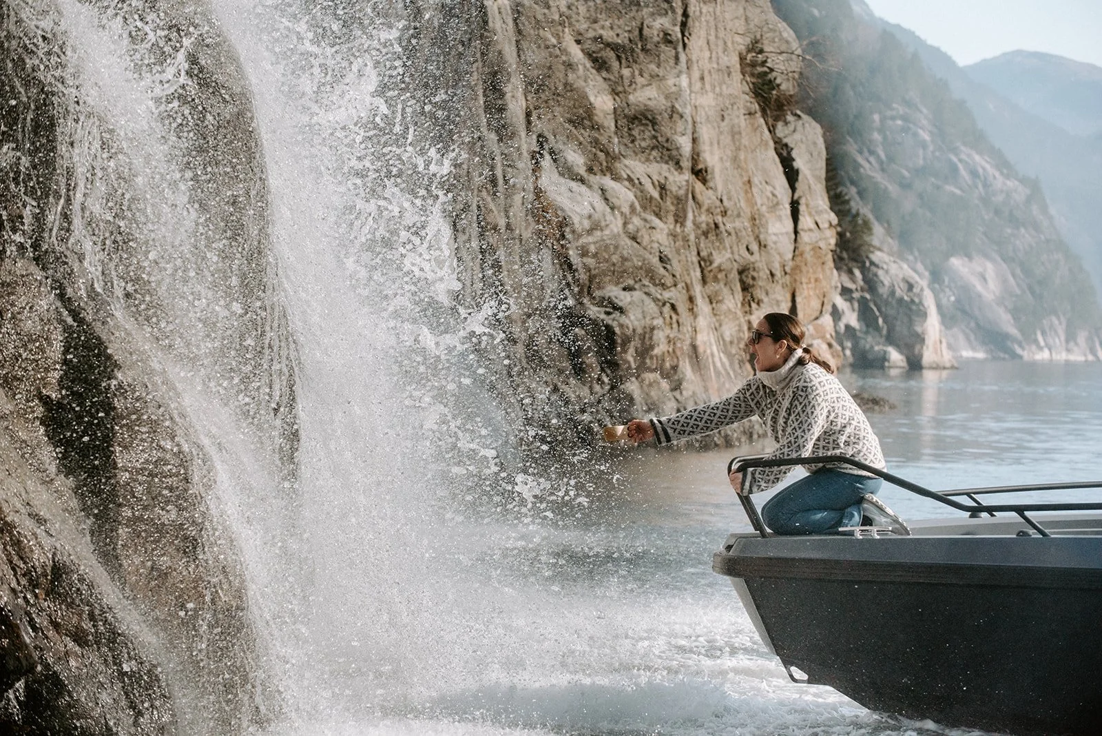 Guest on a Lysefjorden fjord safari reaching from the boat to fill a cup with fresh mountain water from a waterfall cascading down the cliff.