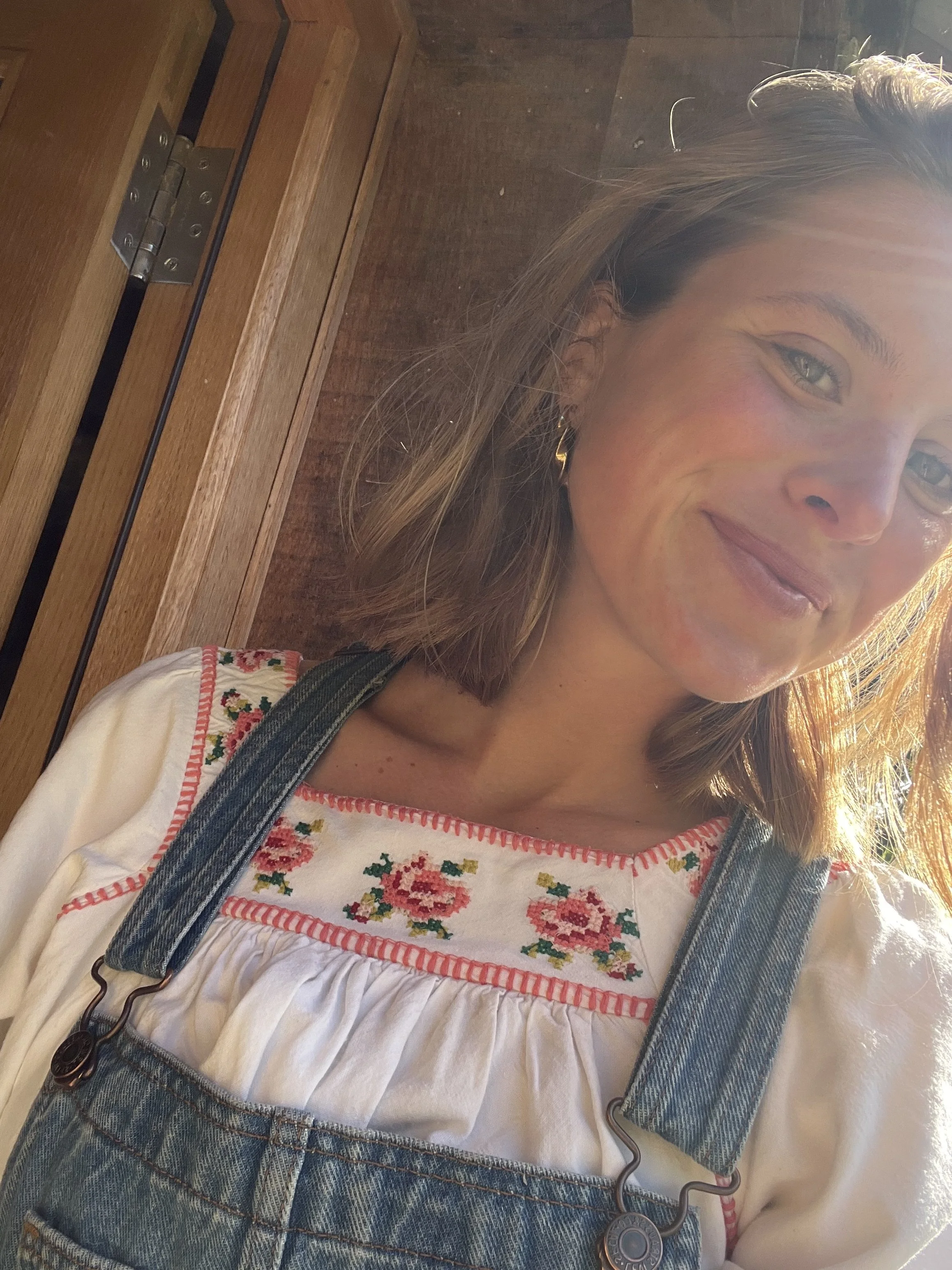 A young woman with light brown hair, green eyes, and earrings smiling, wearing a white embroidered blouse with floral patterns and denim overalls, in front of a wooden background.