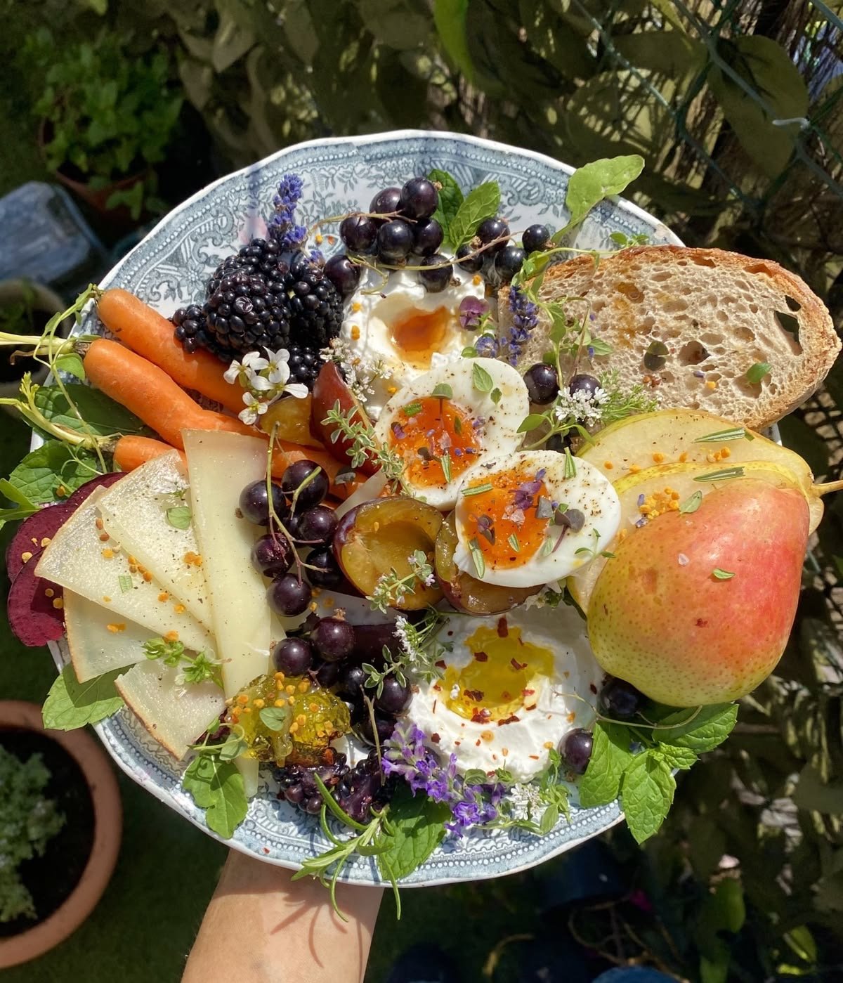 A plate of breakfast with soft-boiled eggs, fresh fruit, cheese, bread, and berries, garnished with herbs and flowers.