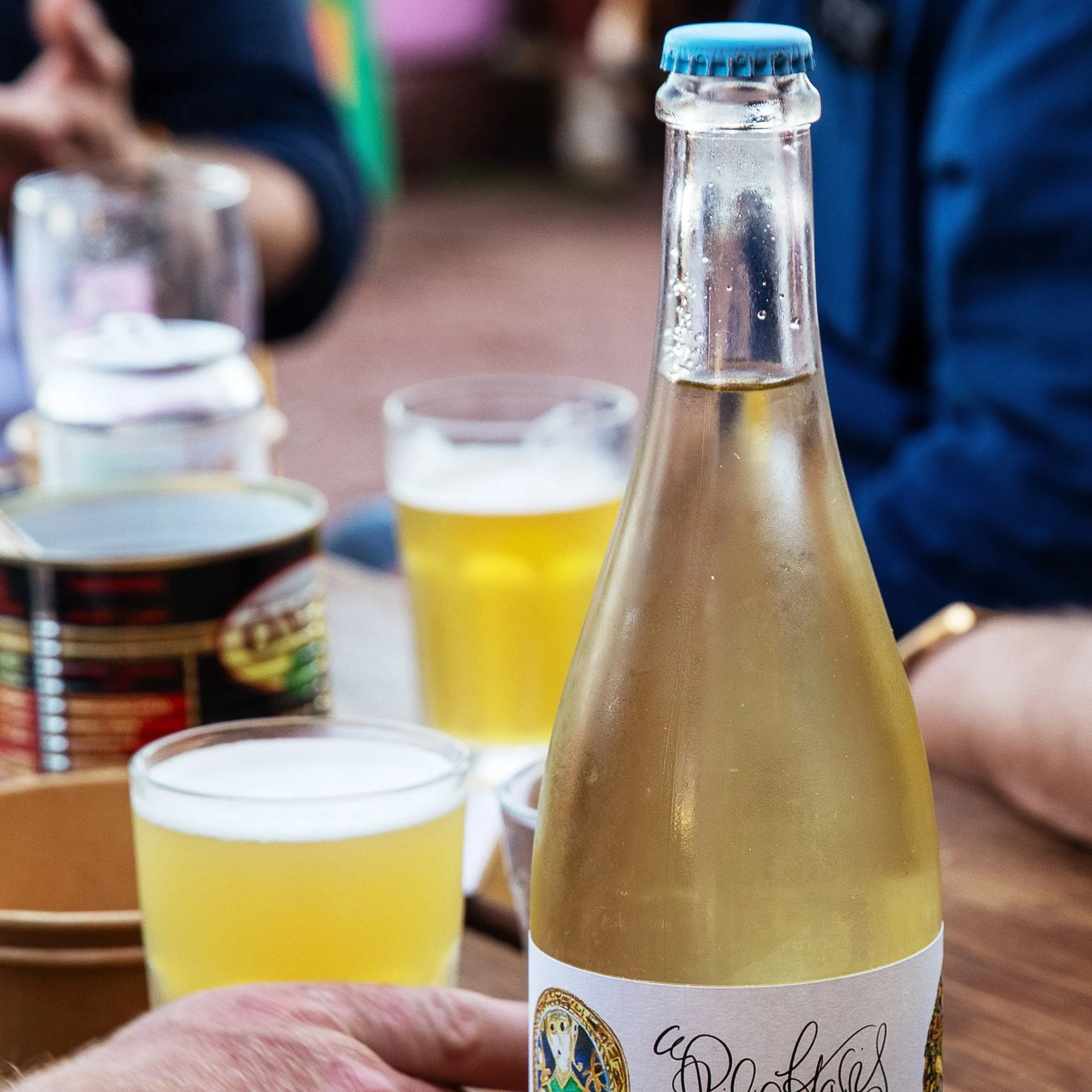 Close-up of a clear glass bottle with a blue cap, containing a light-colored beverage, on a table with blurred cans and cups in the background.