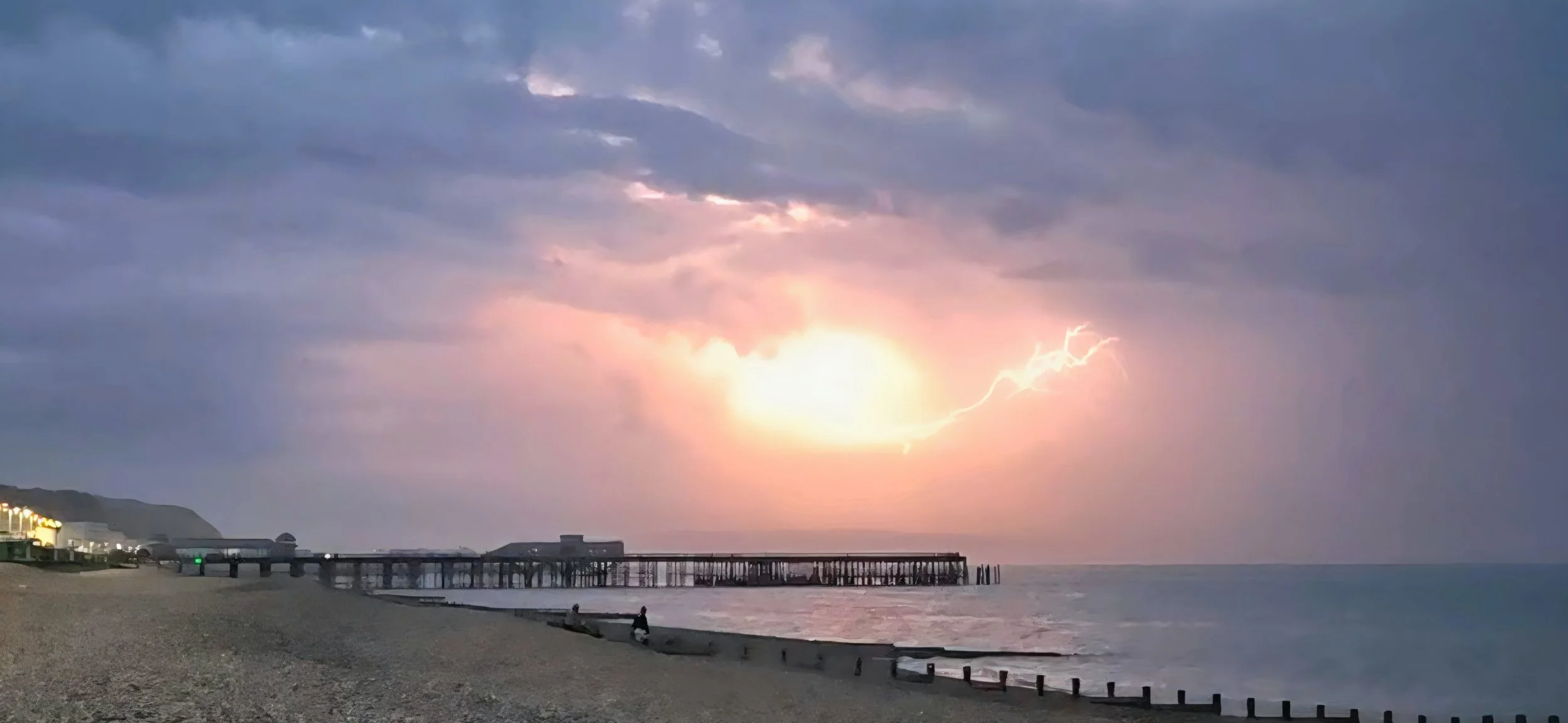A beach scene during a thunderstorm at sunset, with a lightning bolt illuminating the dark sky, a pier stretching into the ocean, and some people and benches along the shoreline.