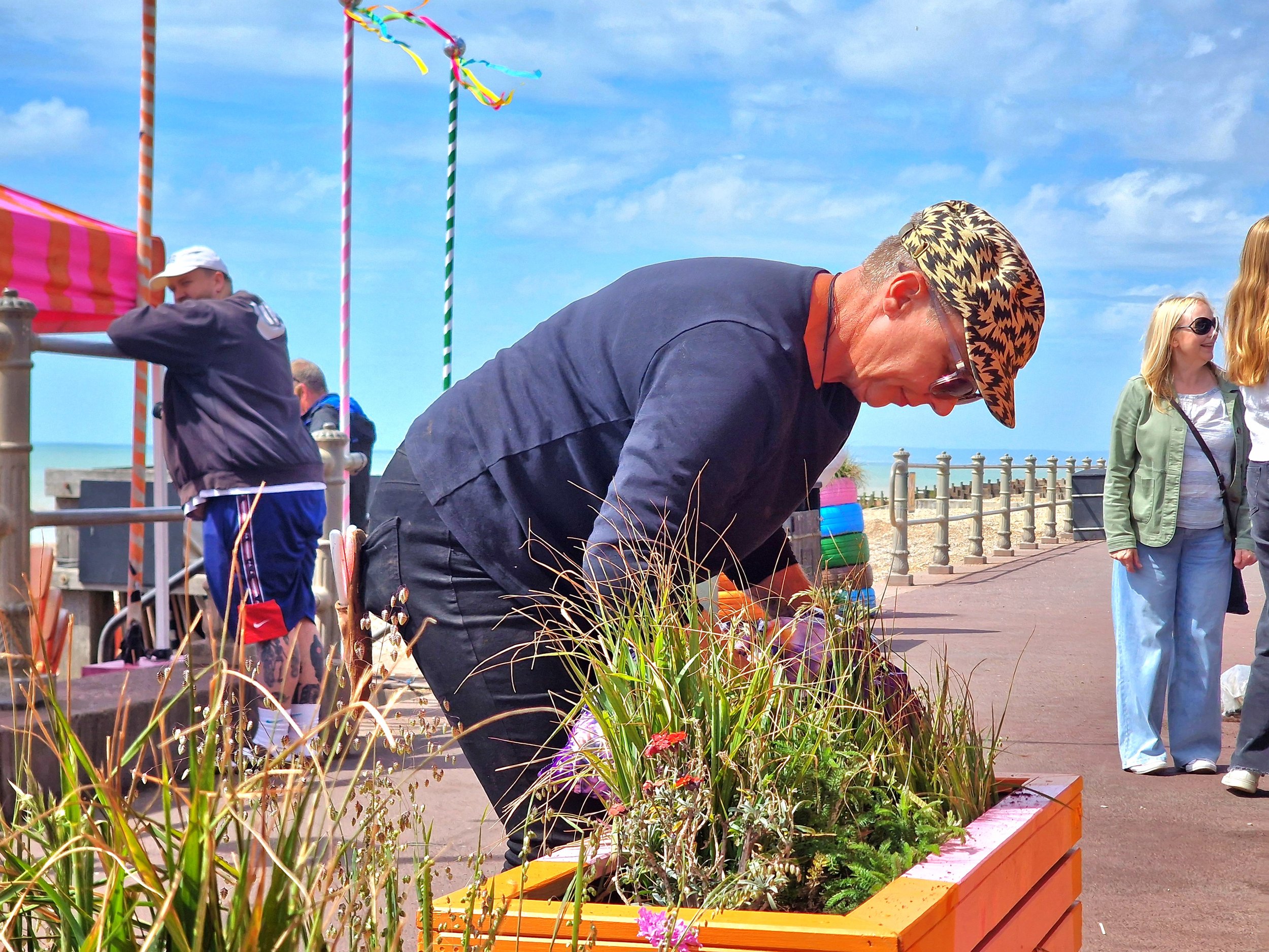 People gathering outdoors near a flower bed on a sidewalk, with a bright blue sky and some clouds overhead. A man in a patterned hat is tending to the plants, while others stand nearby smiling and talking.