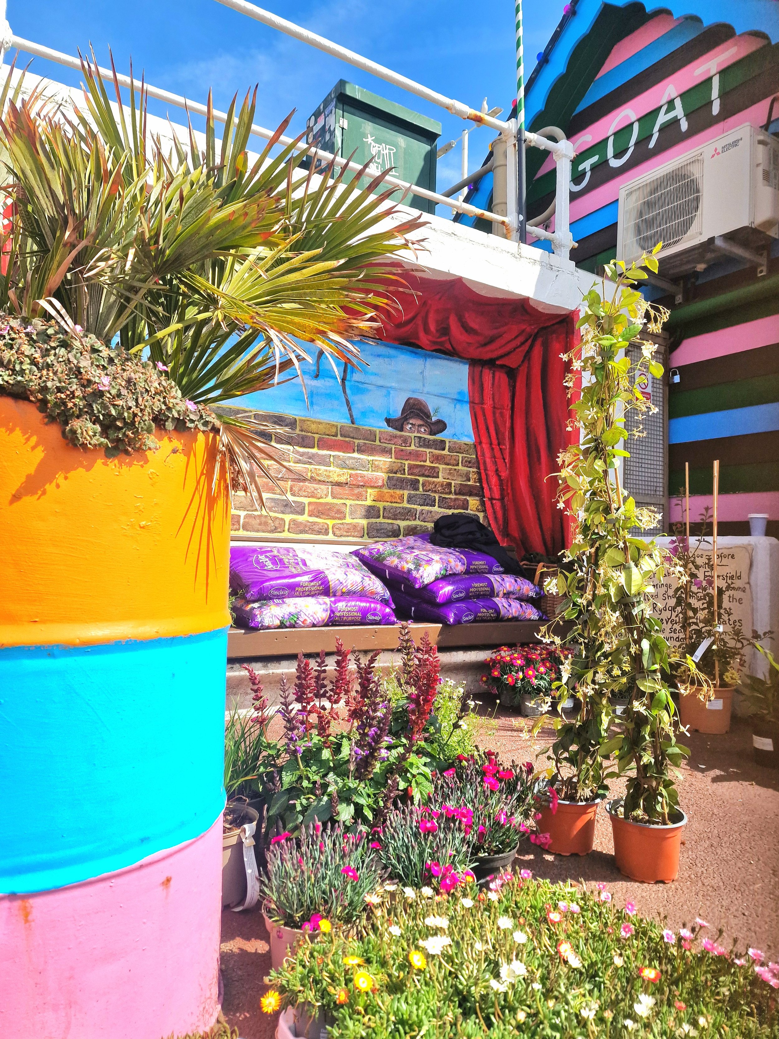 Colorful outdoor market or garden with large painted barrel in pink, blue, and yellow, filled with plants and flowers. Potted plants and flowers are arranged on the ground, with a mural or painted wall featuring a stage and a person wearing glasses a