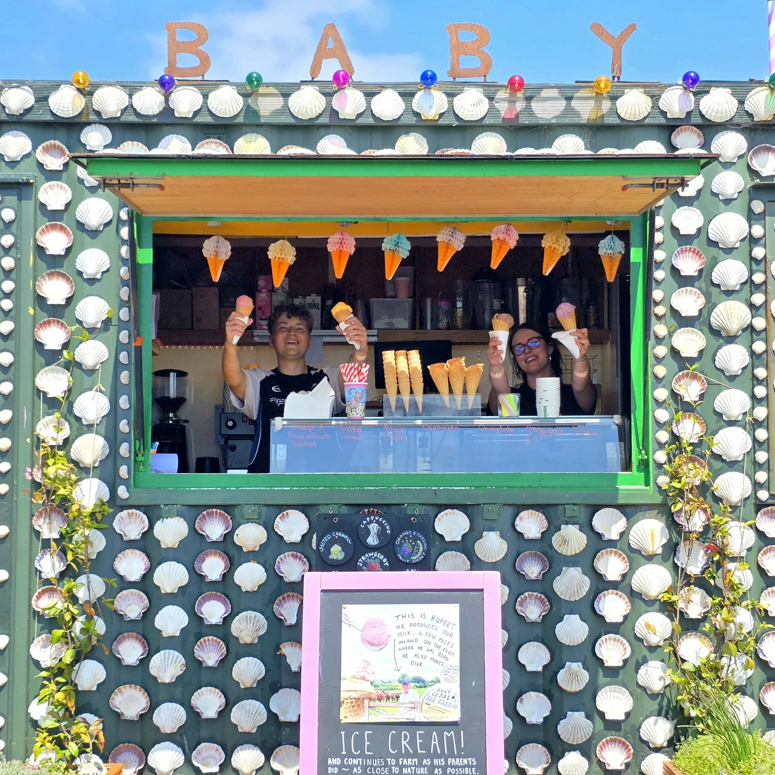 A colorful ice cream stand decorated with seashells and balloons, with two children smiling and holding ice cream cones.