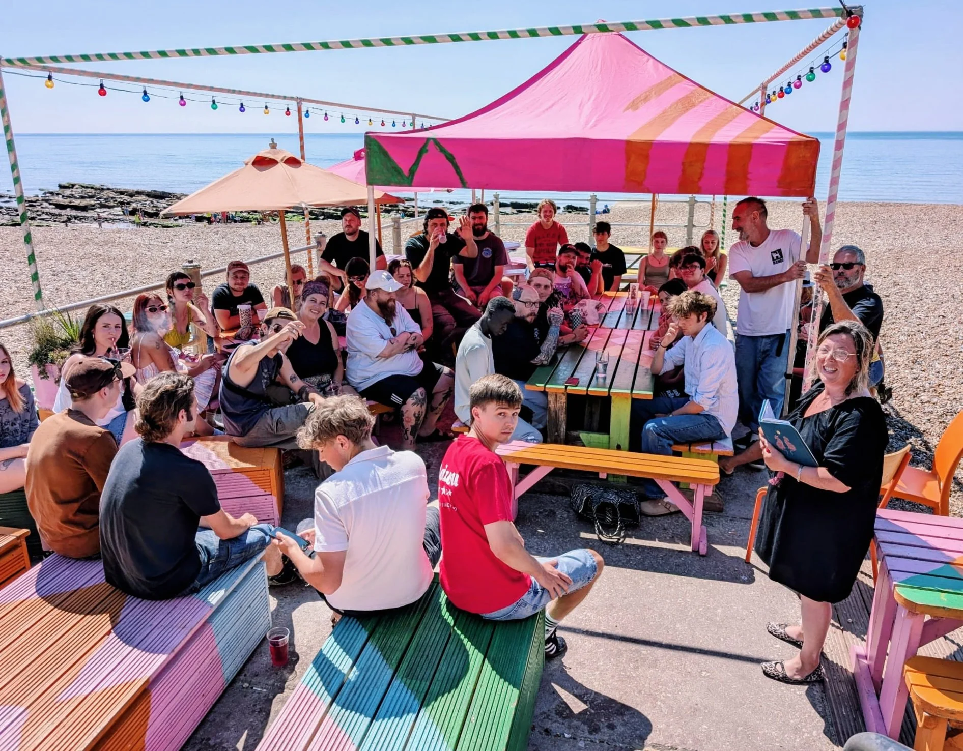 A large group of people gathered under colorful canopies and string lights on a beach, enjoying a sunny day. Some people are sitting, others are standing, and a few are smiling or talking.