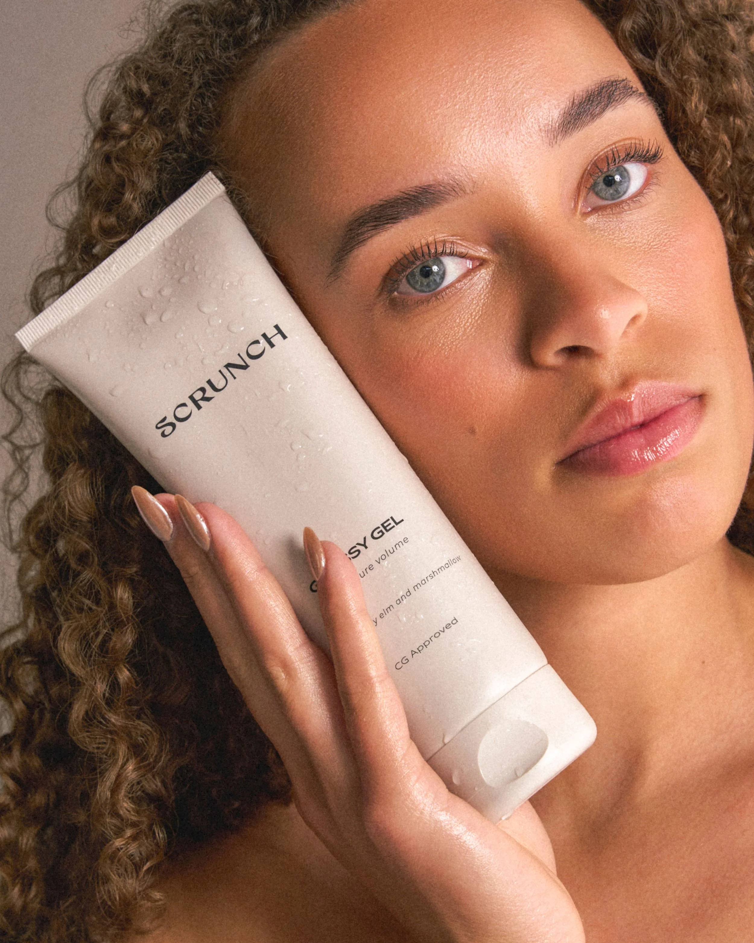 A close-up lifestyle shot of a woman with curly brown hair holding a white tube of SCRUNCH Cosmetics Hydration Scrunch Gel next to her face.