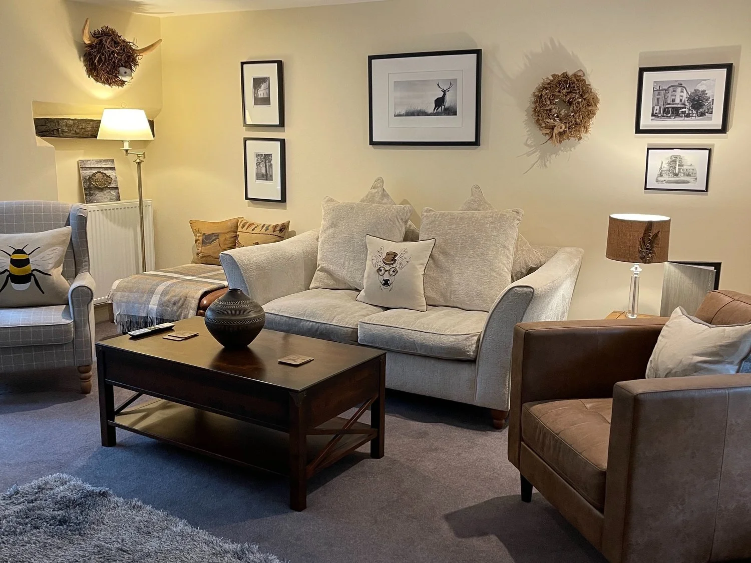 Cozy living room with beige sofa, gray armchair, and brown leather chair. Coffee table with decorative vase, framed pictures on walls, and wall-mounted animal head decoration. Floor lamp and patterned cushions add to the decor.