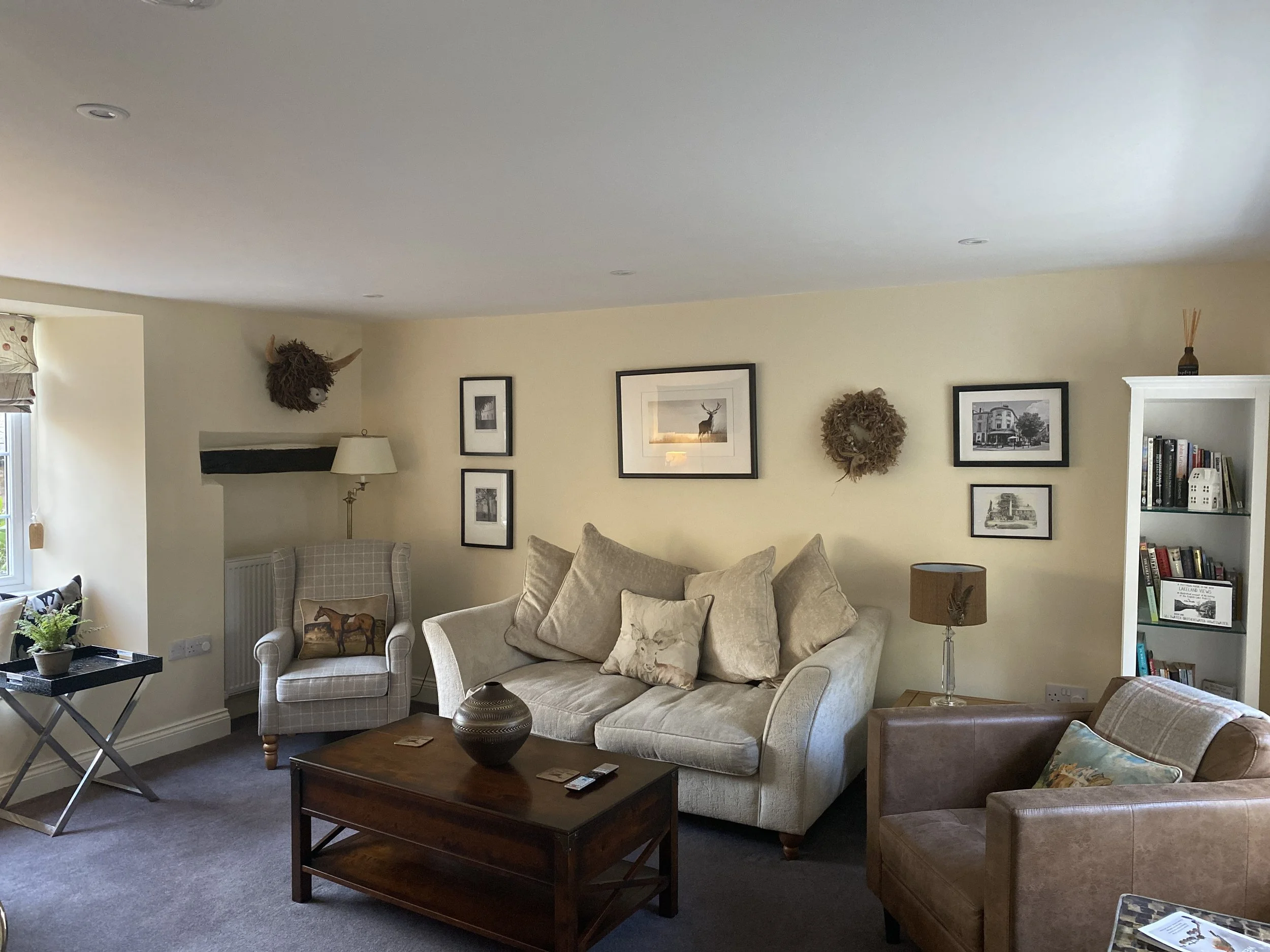Cozy living room with beige sofa, armchairs, and coffee table; wall art and decorative pieces; bookshelves with books. A window and potted plant are visible.