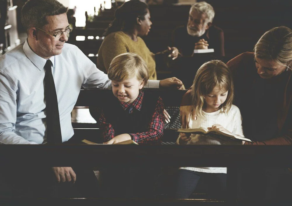 Family sitting together in church pews while two children read from books during a church service.
