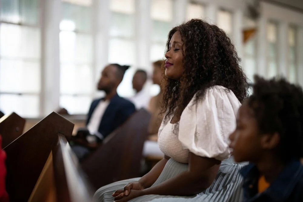Woman sitting in a church pew with eyes closed during a worship service, reflecting peace, prayer and spiritual focus.