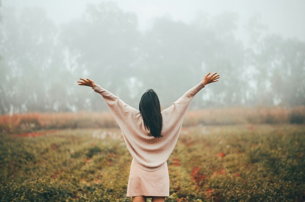 Back view of a woman standing in a field with her arms raised wide toward a foggy forest.