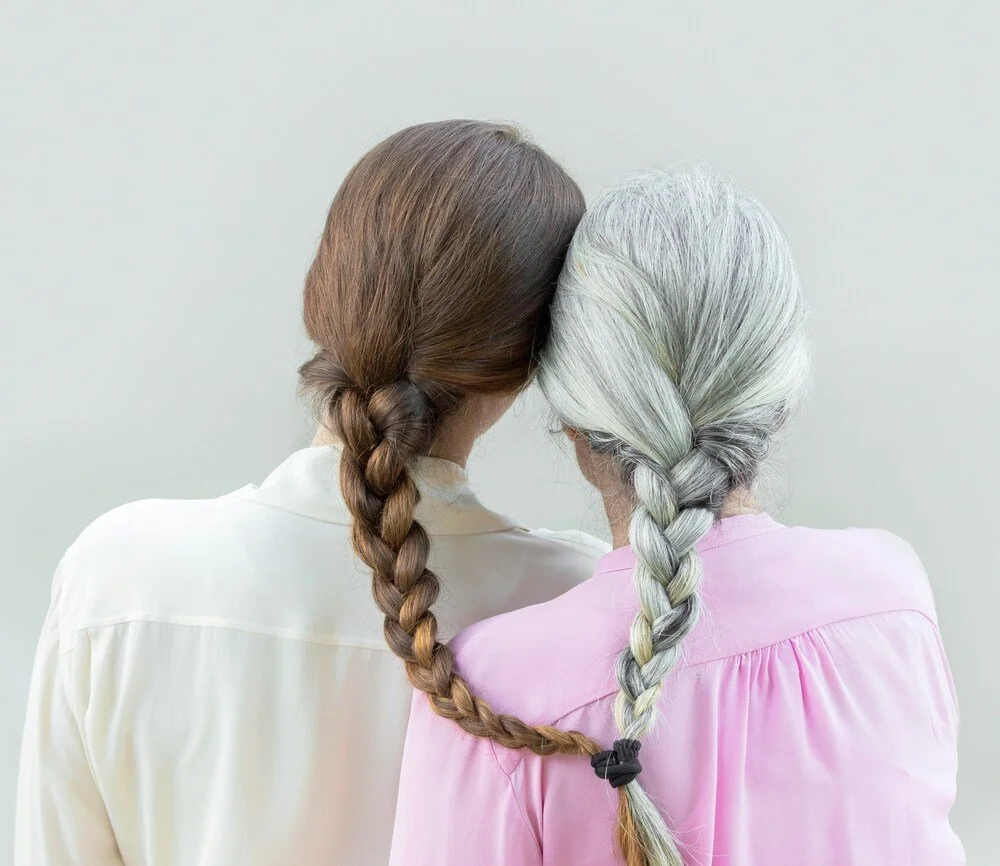 Rear view of a young woman and an older woman with grey hair sitting side by side, their braided hair intertwined.