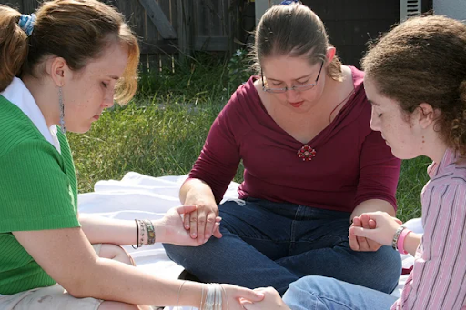 Three people sitting together outdoors holding hands in prayer, representing faith, support, and Christian community.