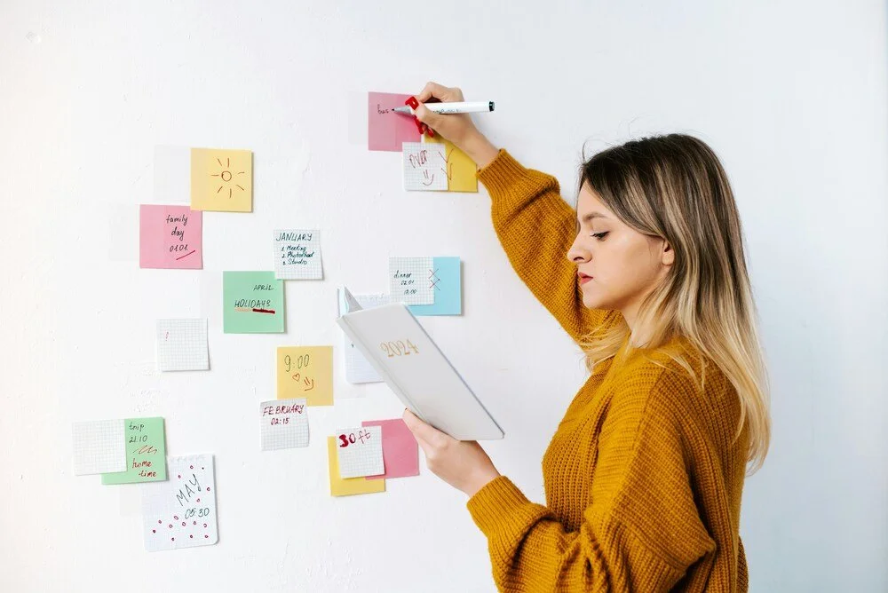 A woman holding a planner and writing on colorful sticky notes on a wall.